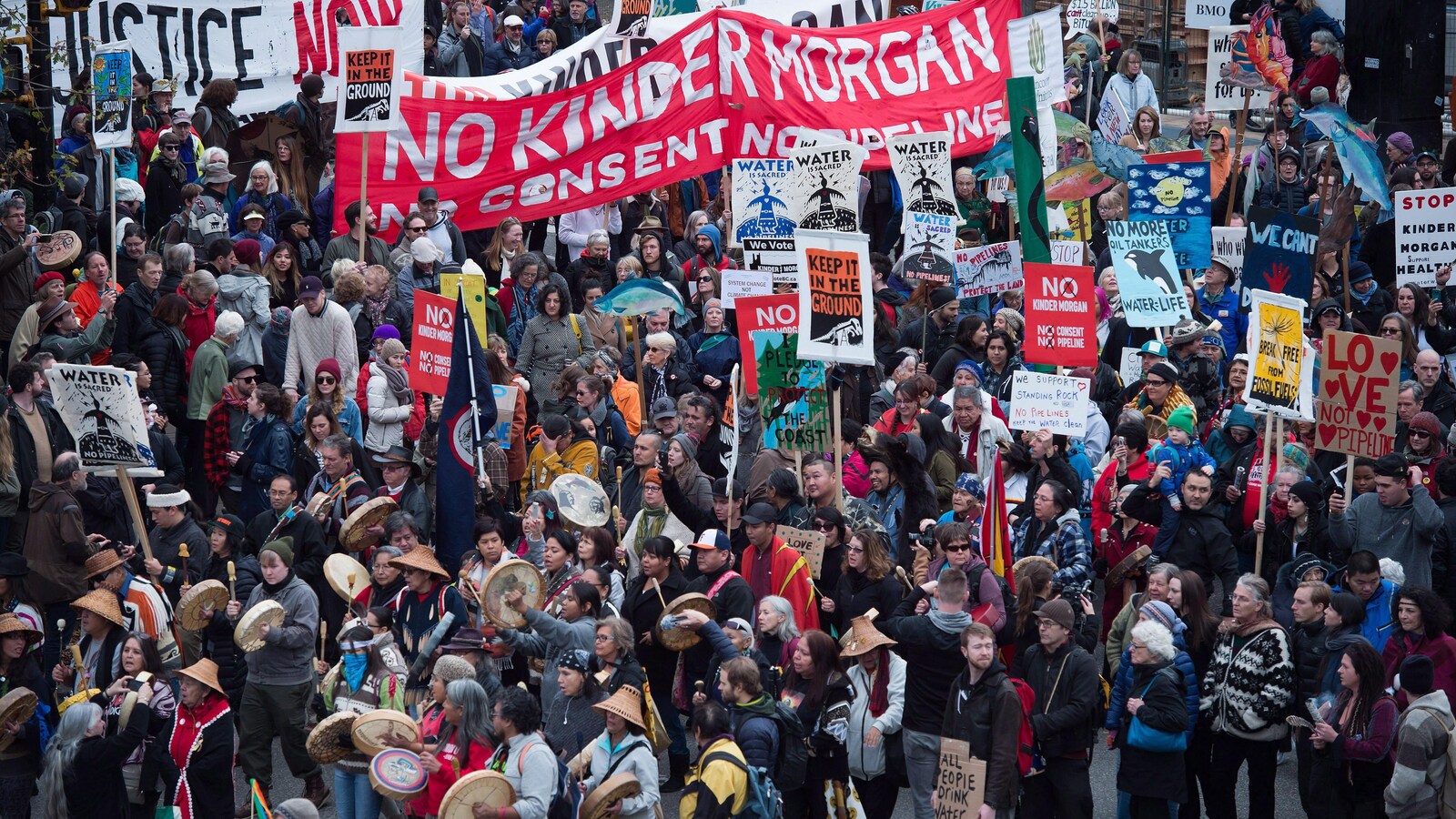 Des milliers de manifestants, certains en habits traditionnels autochtones, et plusieurs qui tiennent des pancartes sur lesquelles on peut lire an anglais Non Kinder Morgan, sont rassemblés.