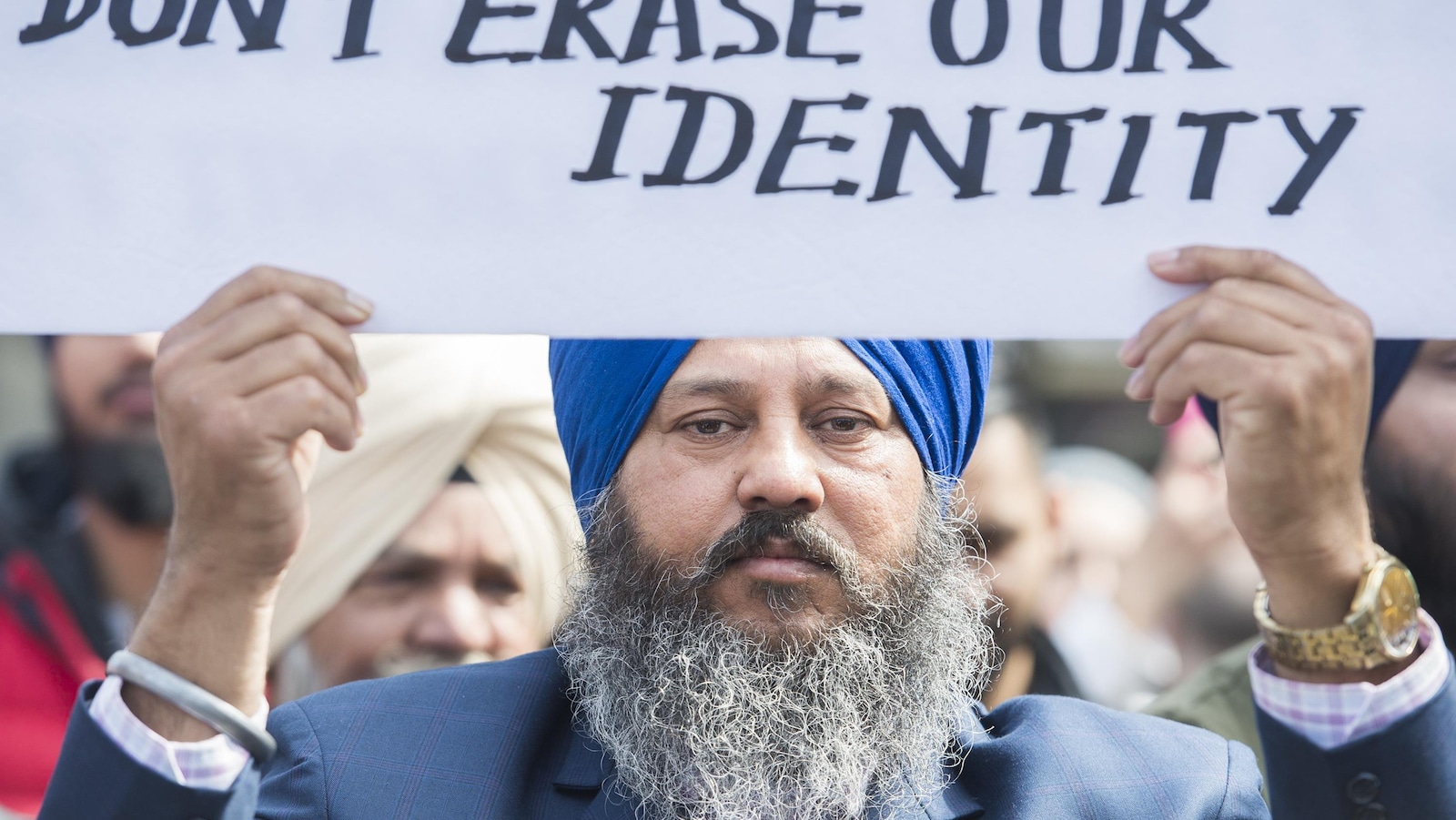 Un homme portant un turban brandit une pancarte disant « Don't erase our identity »