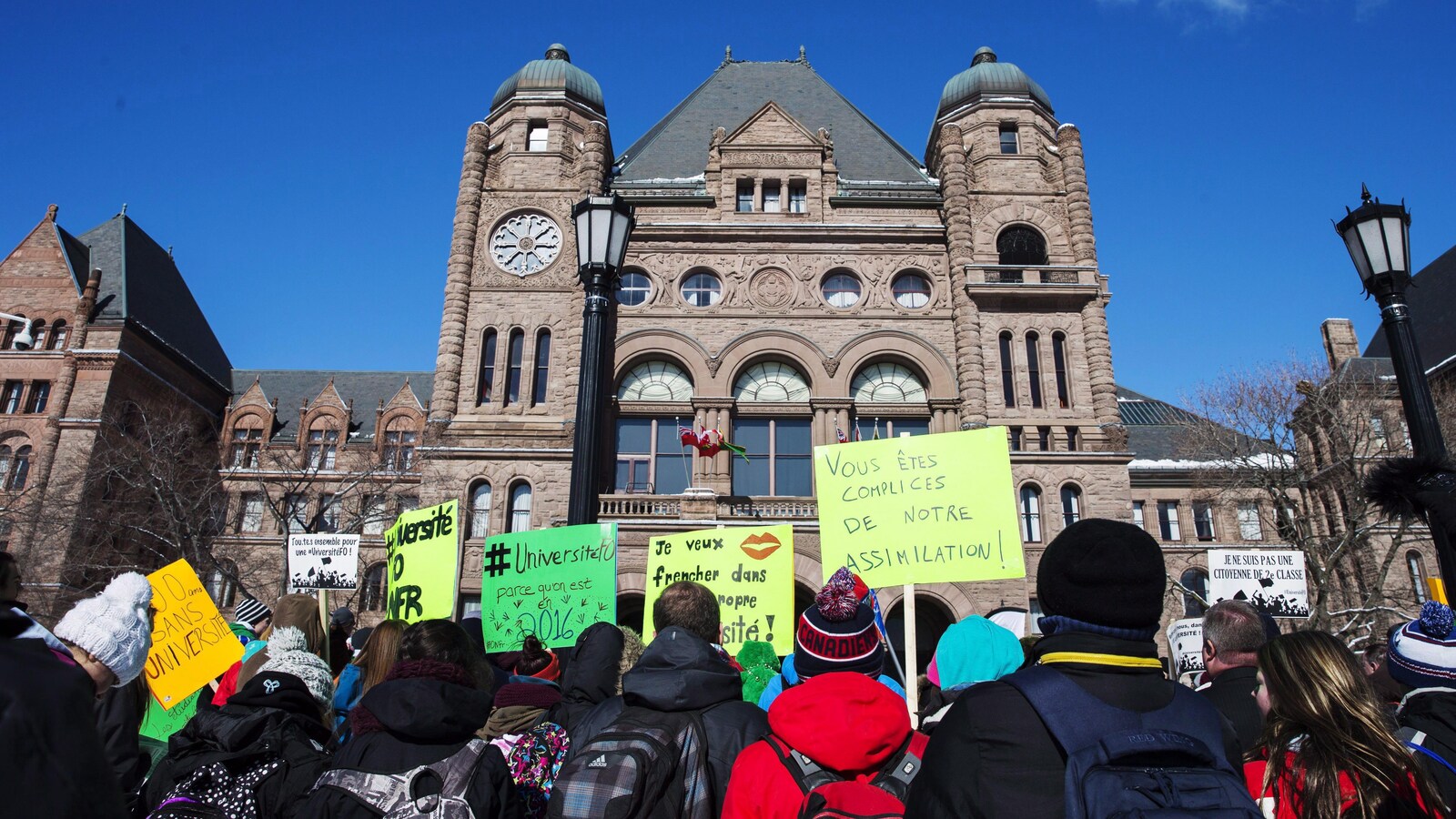 Des manifestants brandissent des pancartes devant l'édifice de l'Assemblée léguislative ontarienne.