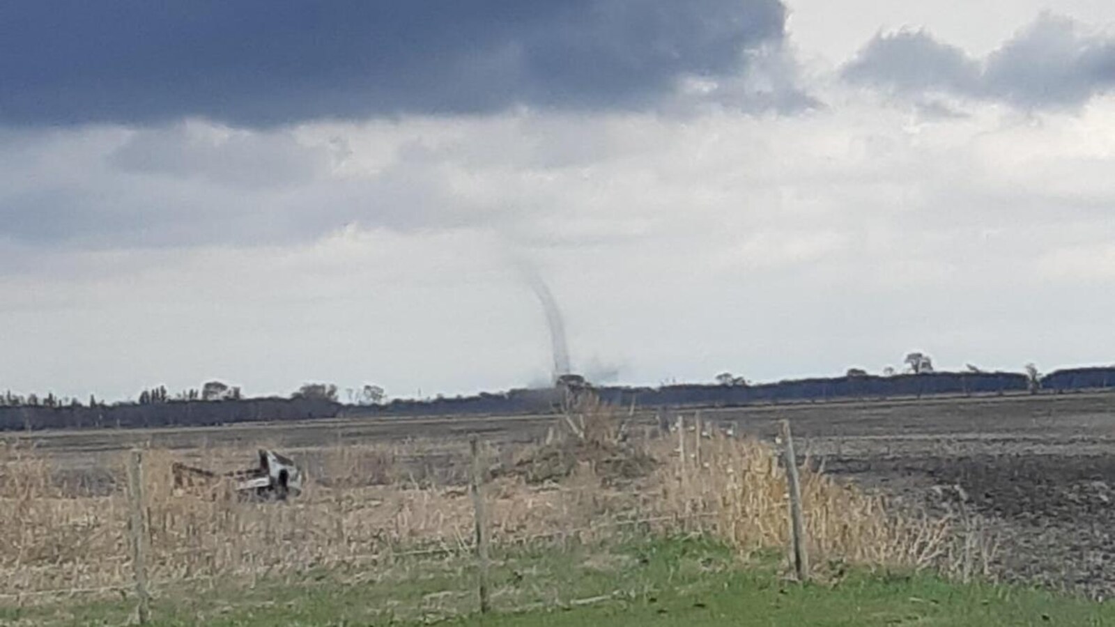 Au milieu d'une terre agricole, une colonne d'air s'élève vers un ciel à moitié blanc et à moitié gris foncé.