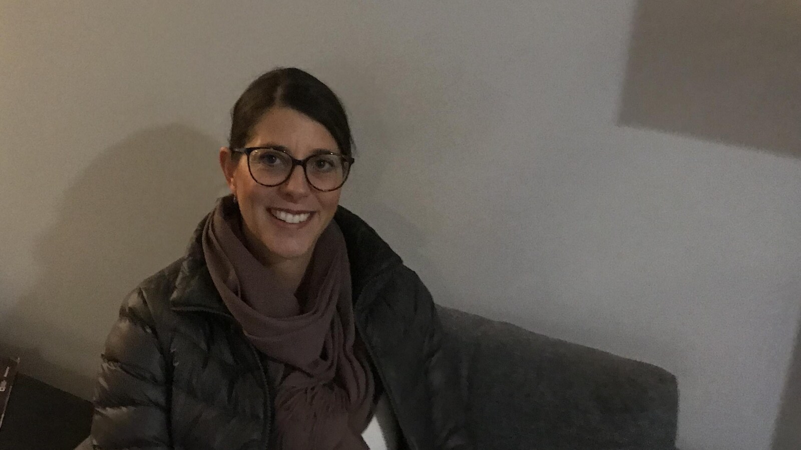 Une femme est assise sur un fauteuil gris devant un mur où est inscrit CBC, Radio-Canada.