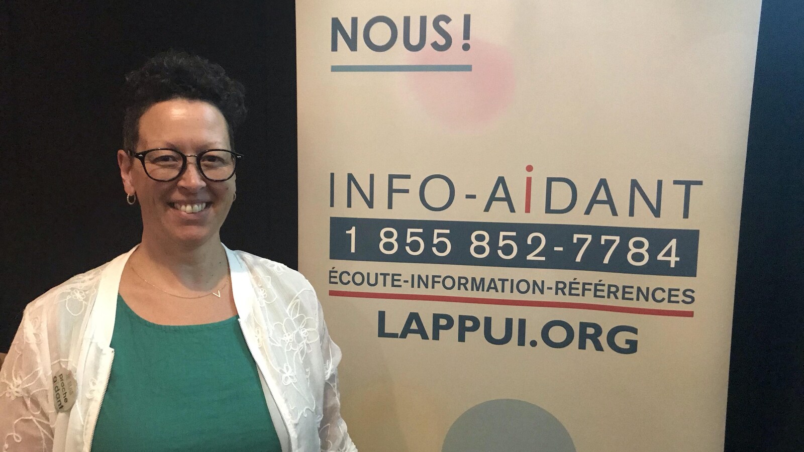 Une femme pose devant une affiche d'Info-Aidant, une ligne téléphonique.