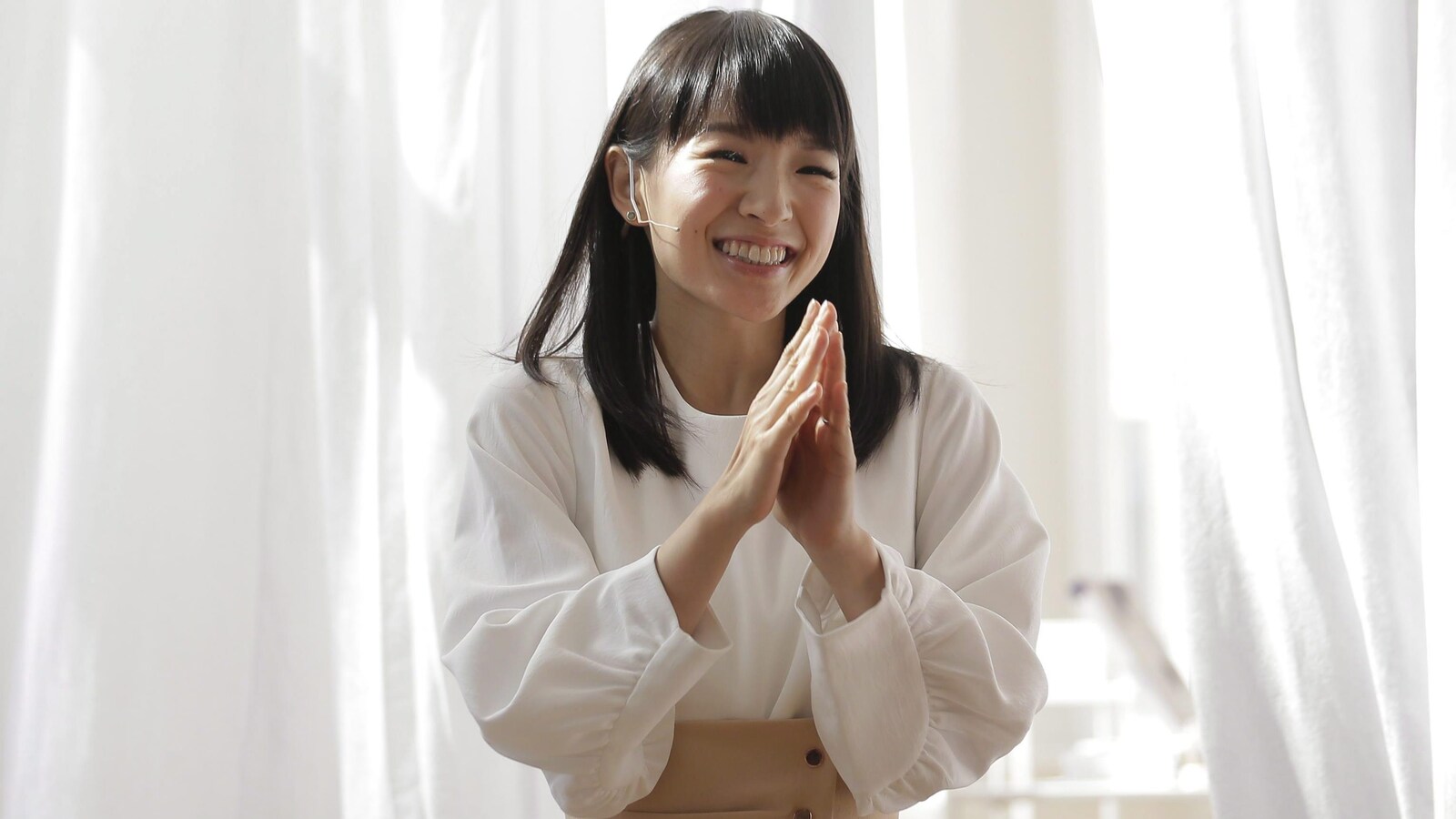 Une japonaise très souriante dans une tunique ample mais serrée à la taille par une large ceinture joint les mains devant des draperies blanches.