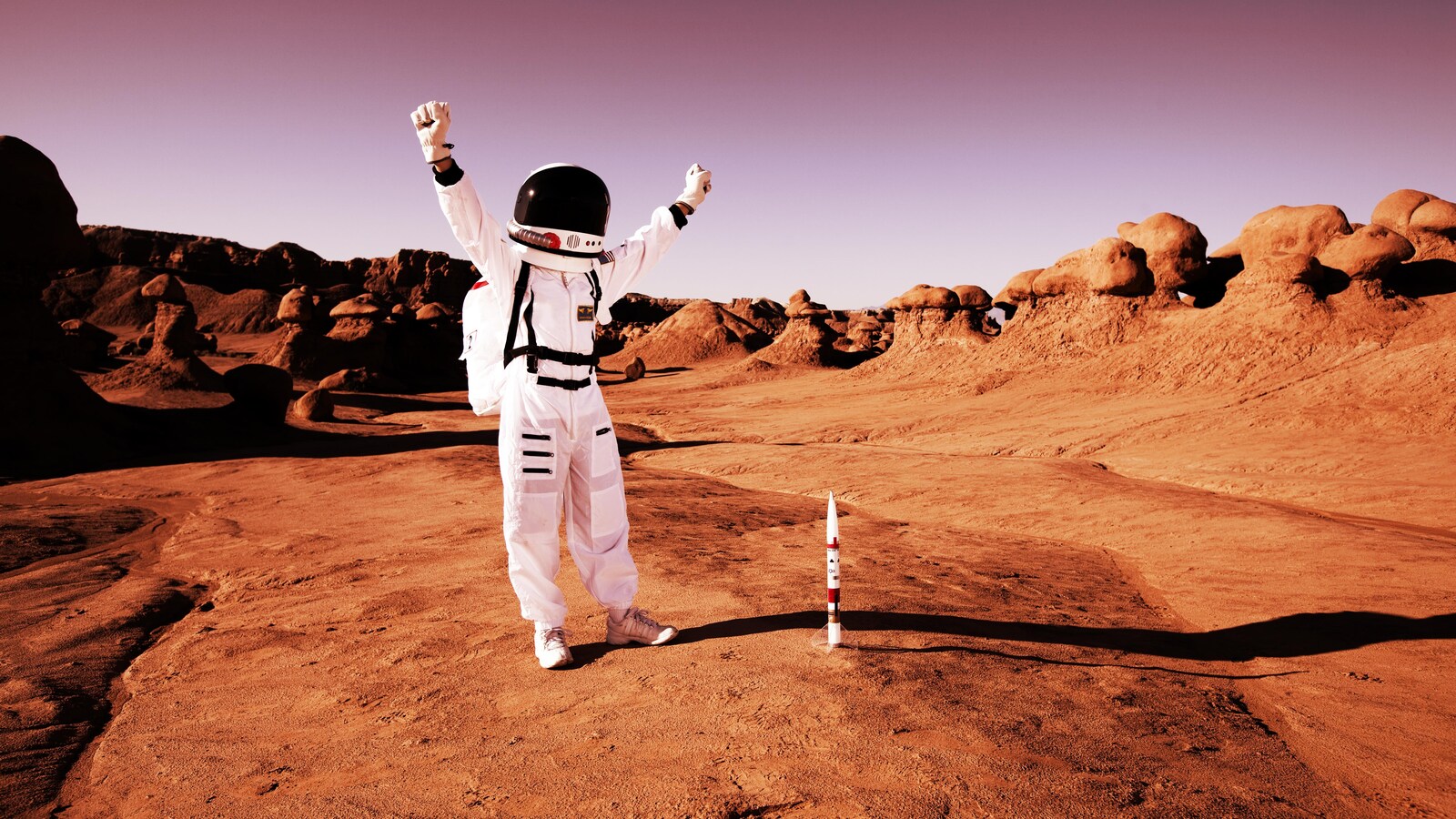 Image représentant un astronaute faisant le signe de la victoire sur une surface rouge ressemblant à celle de la planète Mars.