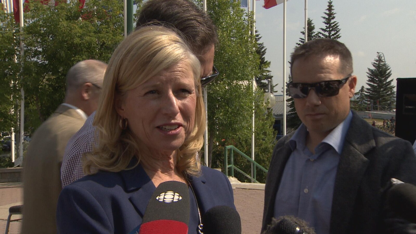 Vue sur une femme avec les cheveux courts et blonds. Elle parle devant des journalistes.