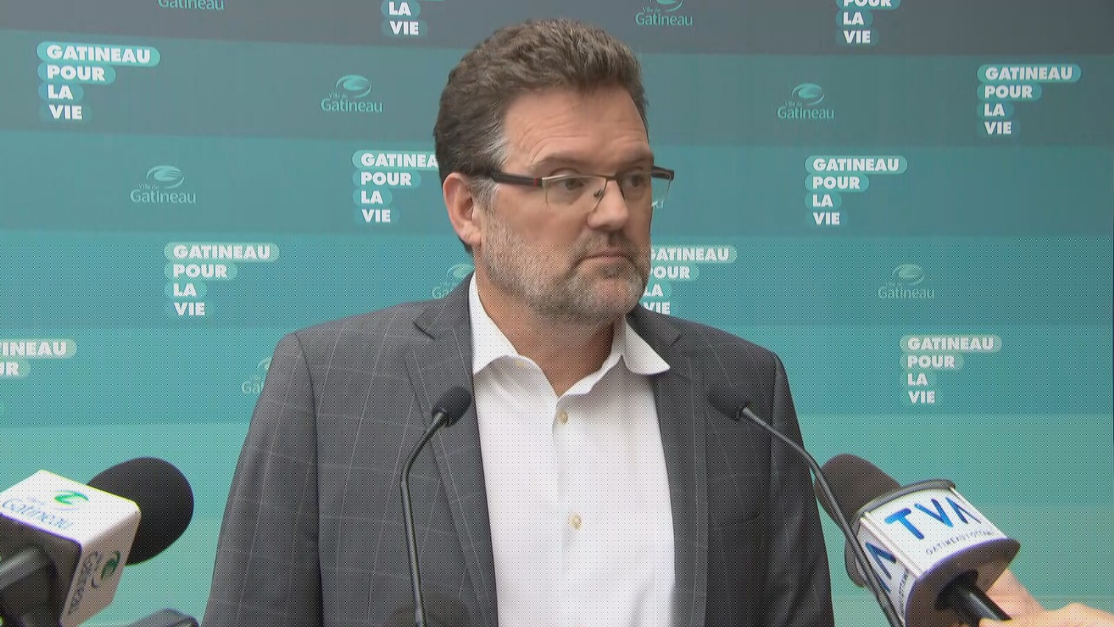 Le maire de Gatineau répond aux questions des journalistes devant une bannière comportant le logo de la Municipalité.