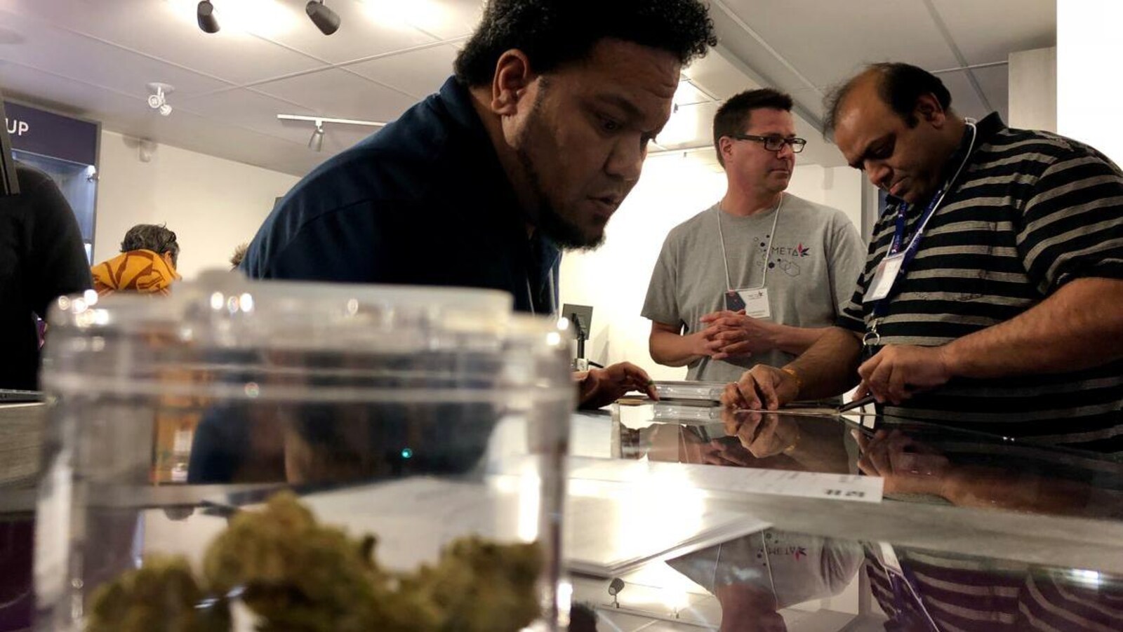 Trois hommes regardent une liste de produits sur un comptoir avec, à l'avant-plan, un pot contenant des cocottes de cannabis.