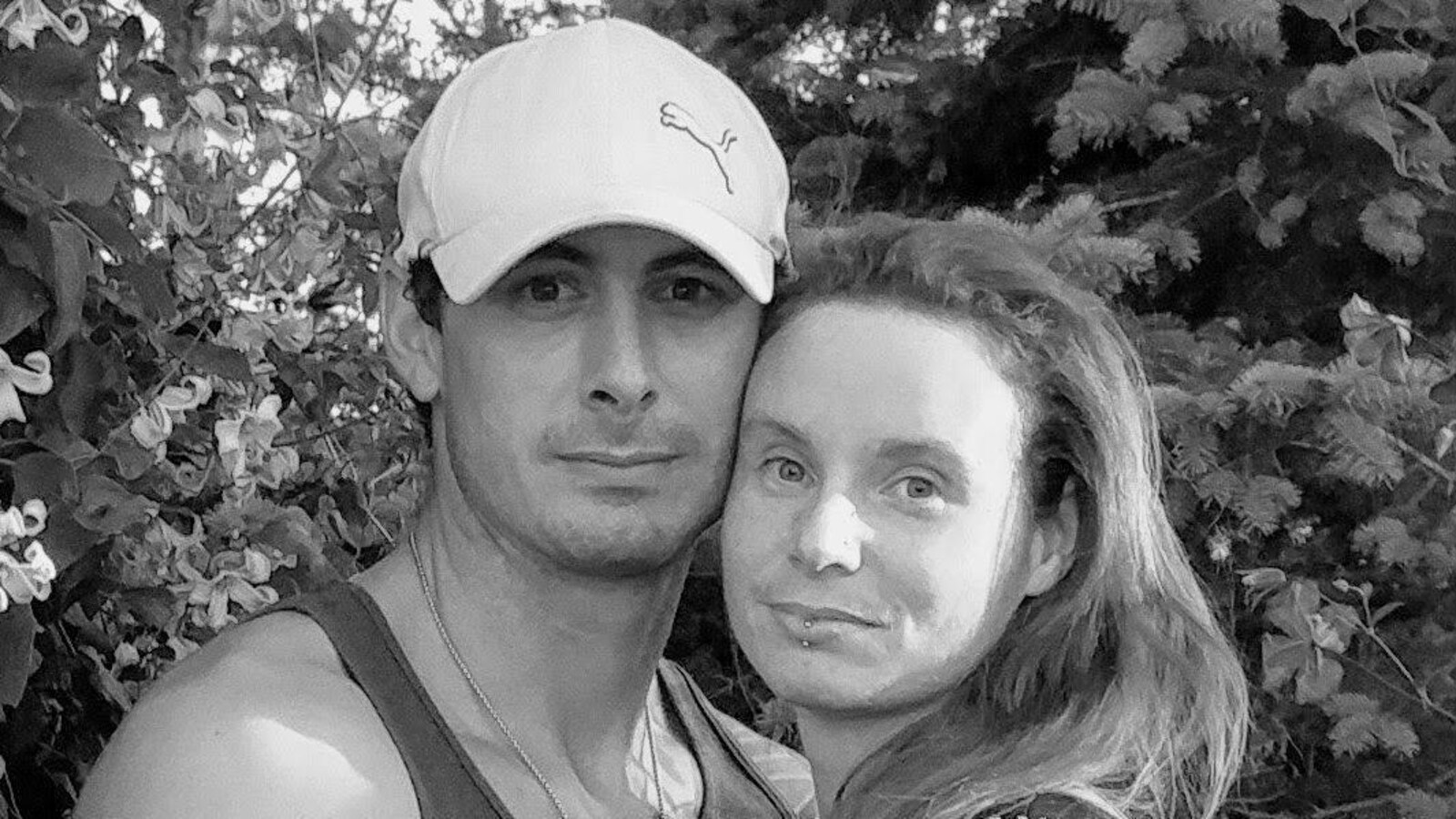 Un jeune couple se tient les bras pour prendre une photo, visages collés, devant des sapins.