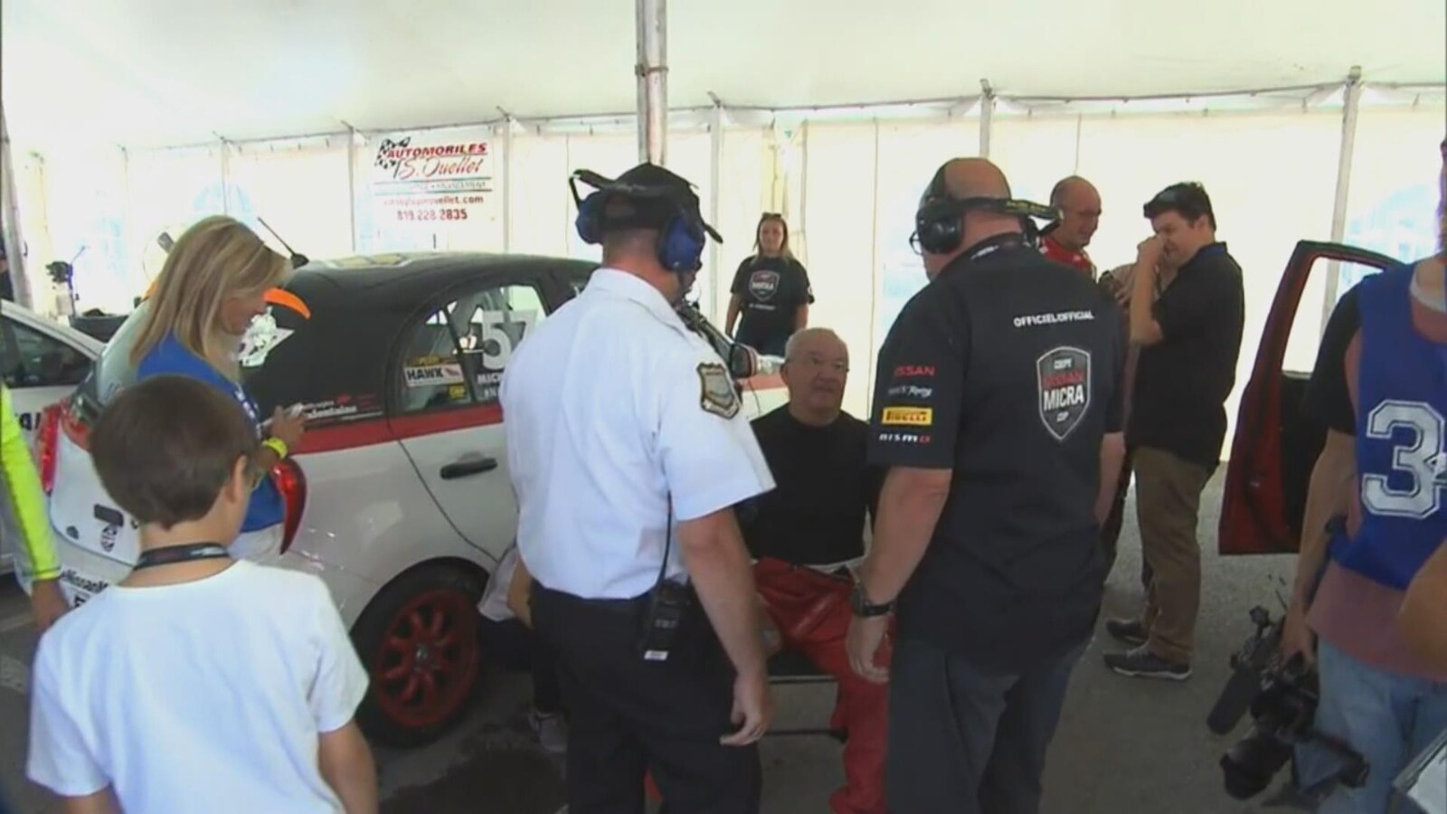 Des hommes, debout, entourent Michel Barrette et discutent avec lui alors qu'il est assis sur une chaise près de son véhicule de course.