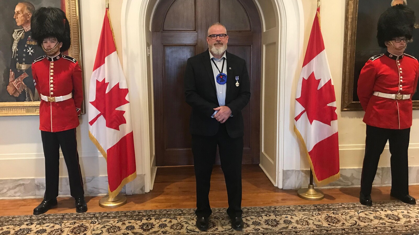 Michel Bourbonnière à Rideau Hall portant sa médaille debout entre deux drapeaux canadiens et deux membres de la Gendarmerie royale du Canada.