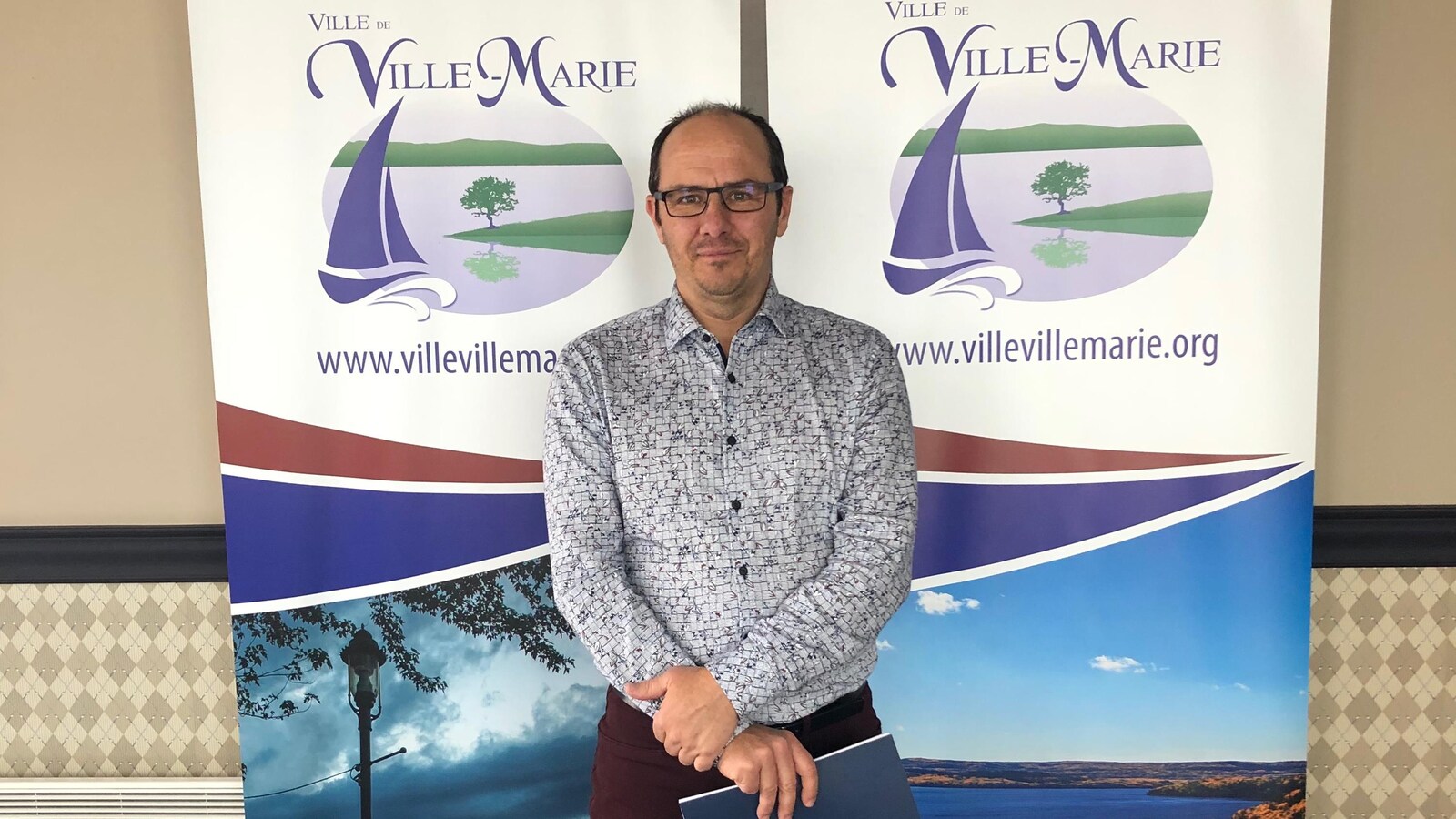 Michel Roy pose devant des affiches représentant Ville-Marie, un document à la main.