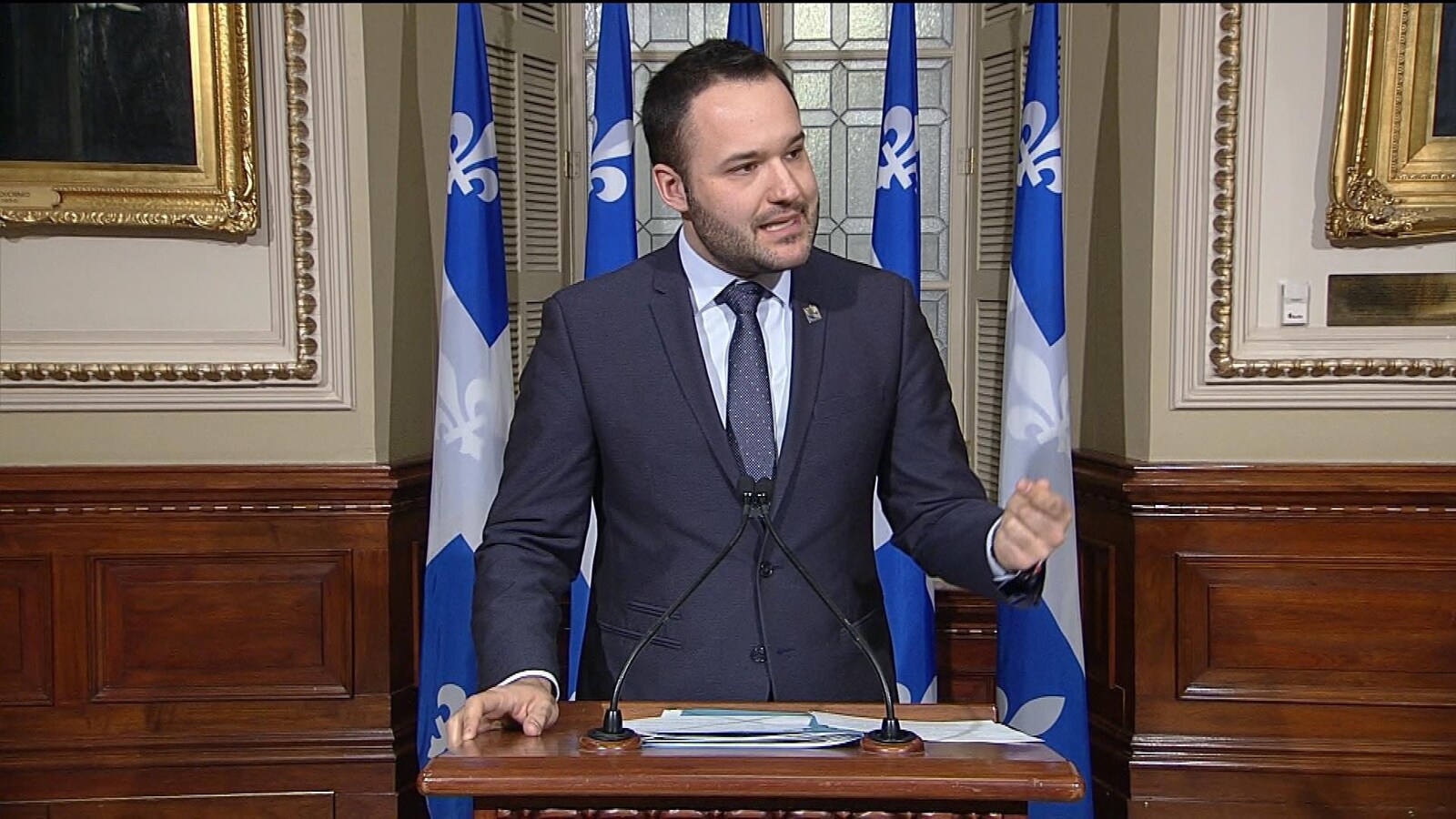 Mathieu Lacombe parle, debout devant des drapeaux du Québec.