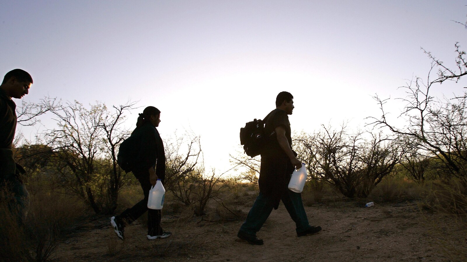 Des migrants marchent dans le désert à l'aube, des cruches d'eau à la main et sacs sur le dos.