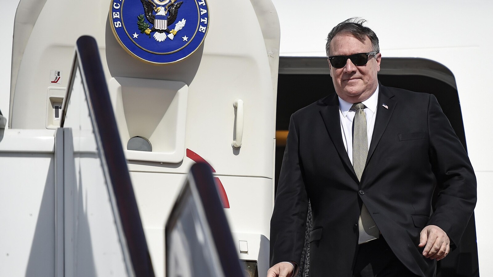 Mike Pompeo, portant un complet avec une cravate et des lunettes de soleil, sort d'un avion.