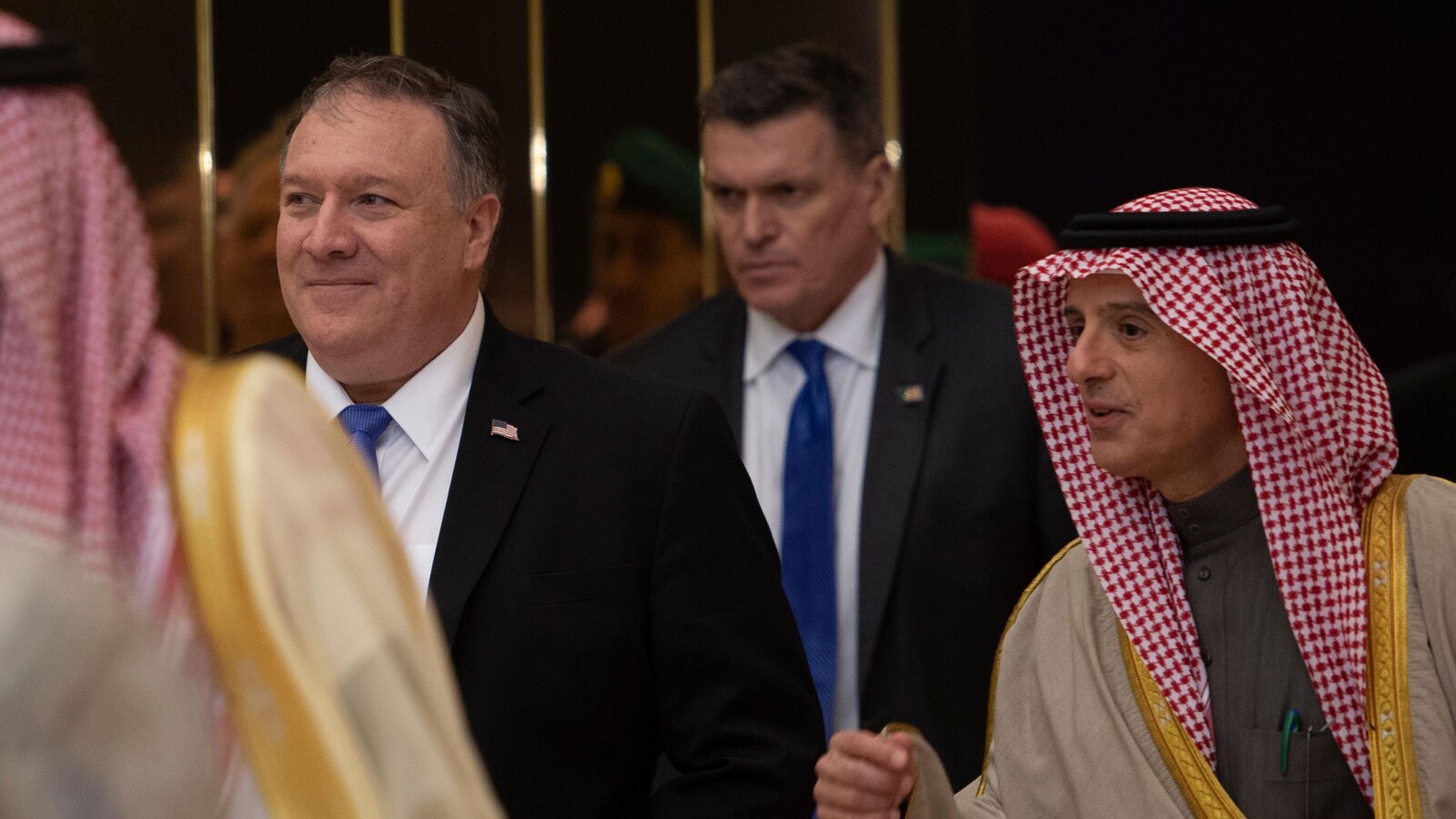 Mike Pompeo, portant un complet et une cravate, marche à côté d'Adel Al-Jubeir qui porte une tenue traditionnelle saoudienne. Ils sont suivi par un agent de sécurité de M. Pompeo.