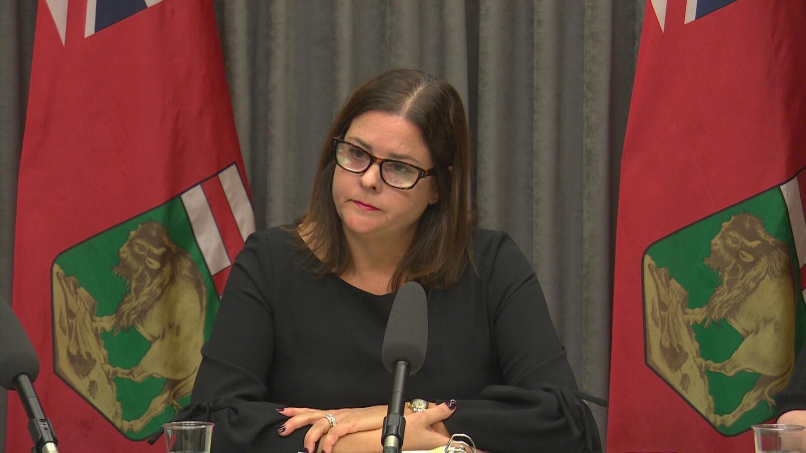 La ministre Stefanson est assise devant deux drapeaux du Manitoba et est habillée de noir.