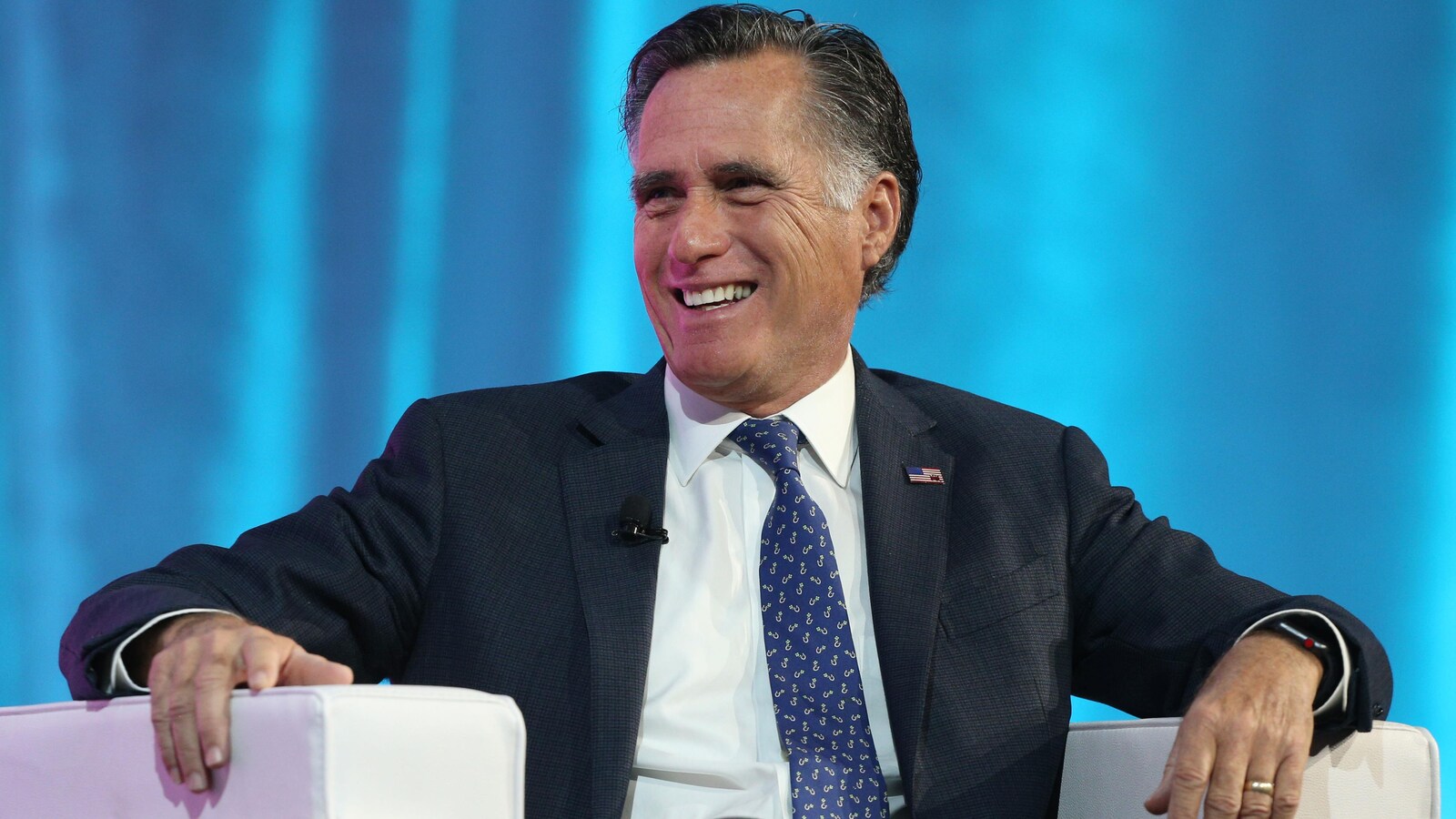 Mitt Romney est assis dans un fauteuil. Il sourit.