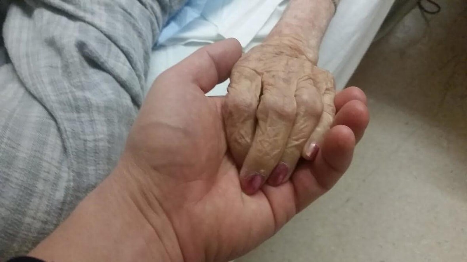 La main d'une femme en fin de vie, allongée sur un lit médical, repose dans la main d'une personne qui semble être en bonne santé. La main de la personne en fin de vie est maigre et ridée, avec les jointures apparentes. Les ongles sont vernis et manucurés.