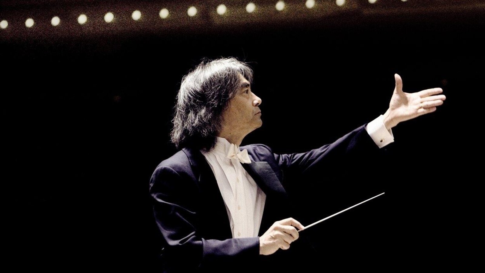 Un chef d'orchestre regarde vers les musiciens, un bras en l'air, et l'autre tenant sa baguette.