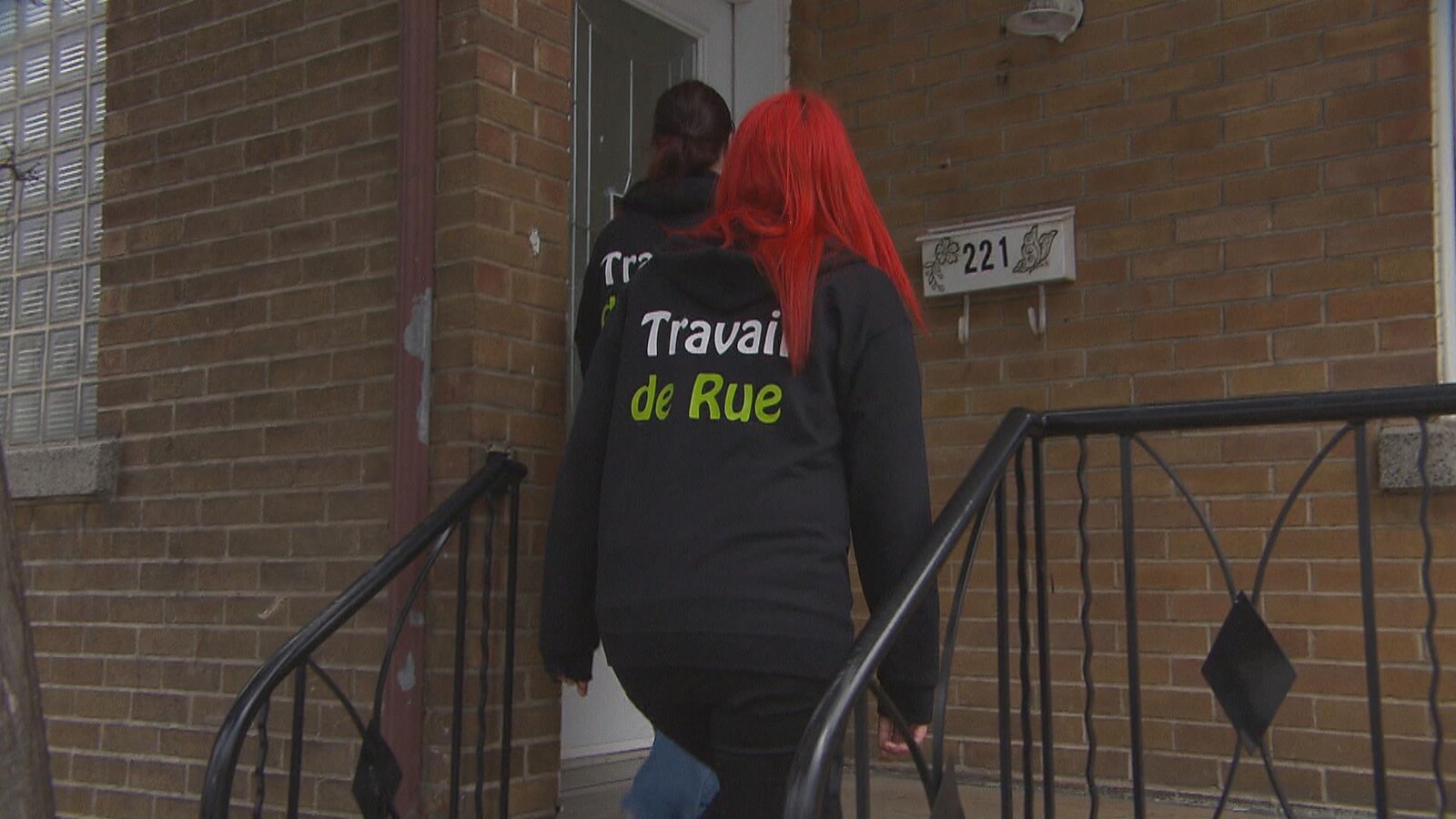 Deux femmes portent un chandail écrit Travail de rue. Elles montent les escaliers d'une maison.