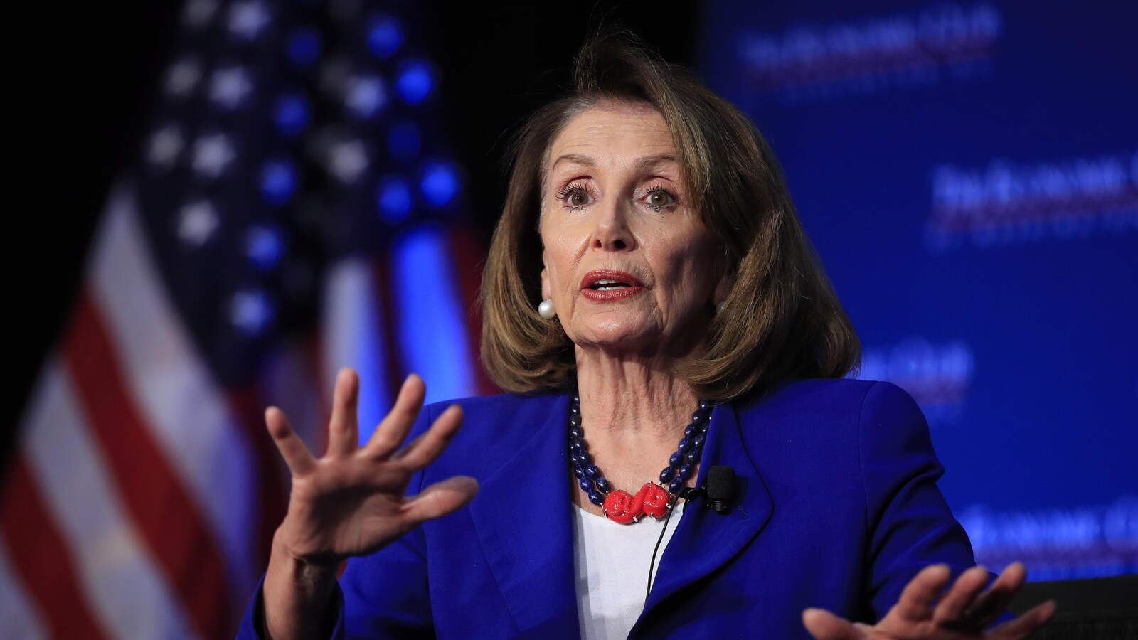 Nancy Pelosi parlant publiquement en bougeant les mains, avec un drapeau américain en arrière-plan.