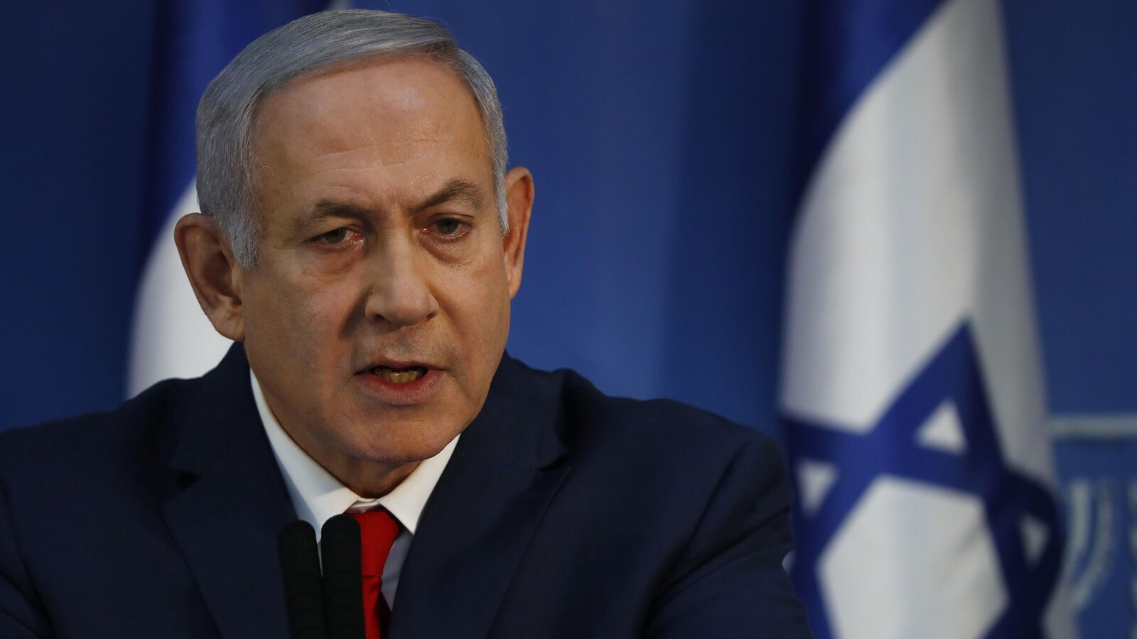 Un homme aux cheveux gris et portant un complet parle derrière un micro et devant des drapeaux d'Israël.