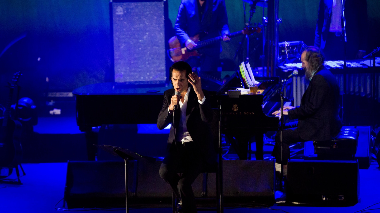 Nick Cave est assis sur un tabouret noir sur la scène éclairée par une lumière bleutée et chante avec son micro dans la main droite.