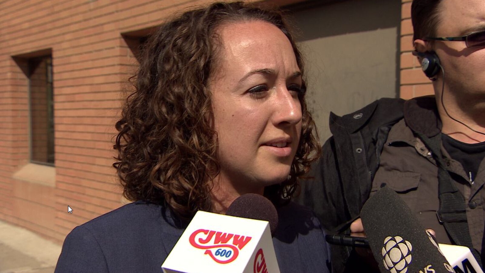 Une femme aux cheveux courts parle à des journalistes devant un bâtiment.