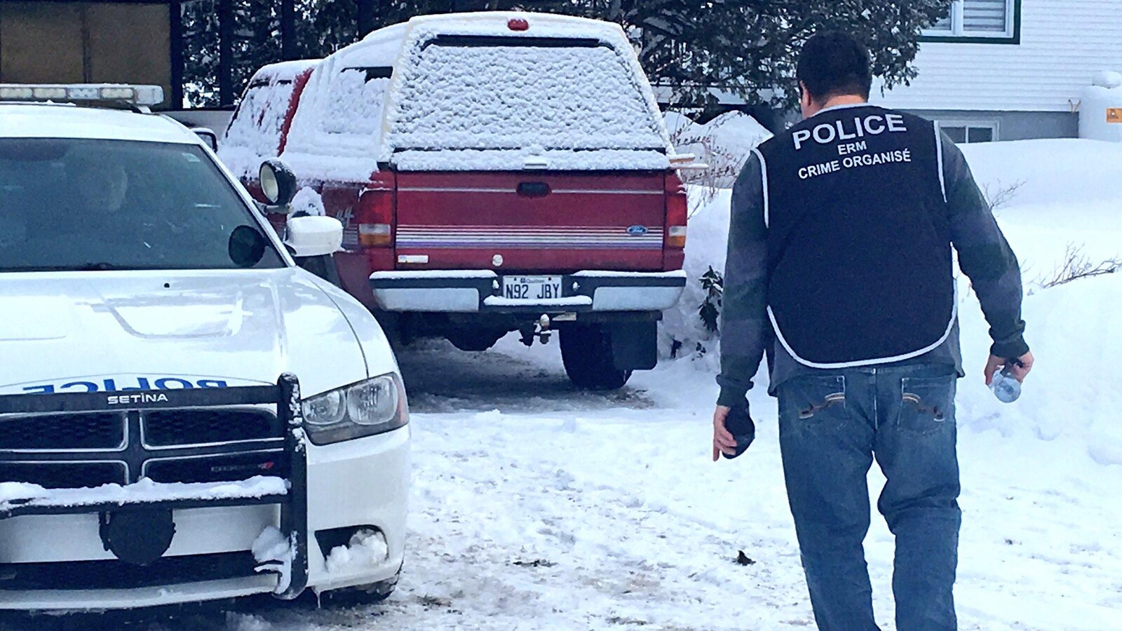 Un policier de l'ERM (crime organisé) se dirige vers une maison. Une voiture de police et un petit camion sont stationnés dans la cour.