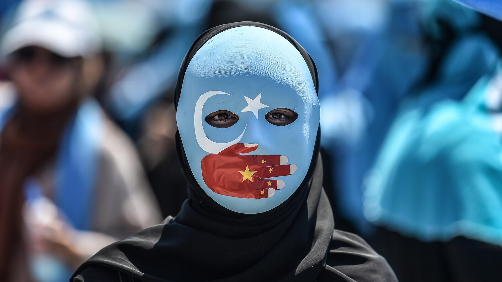 Un manifestant porte un masque à l'effigie du drapeau du Turkestan oriental. Une main représentant le drapeau chinois est peinte et couvre sa bouche. On ne voit que les yeux.