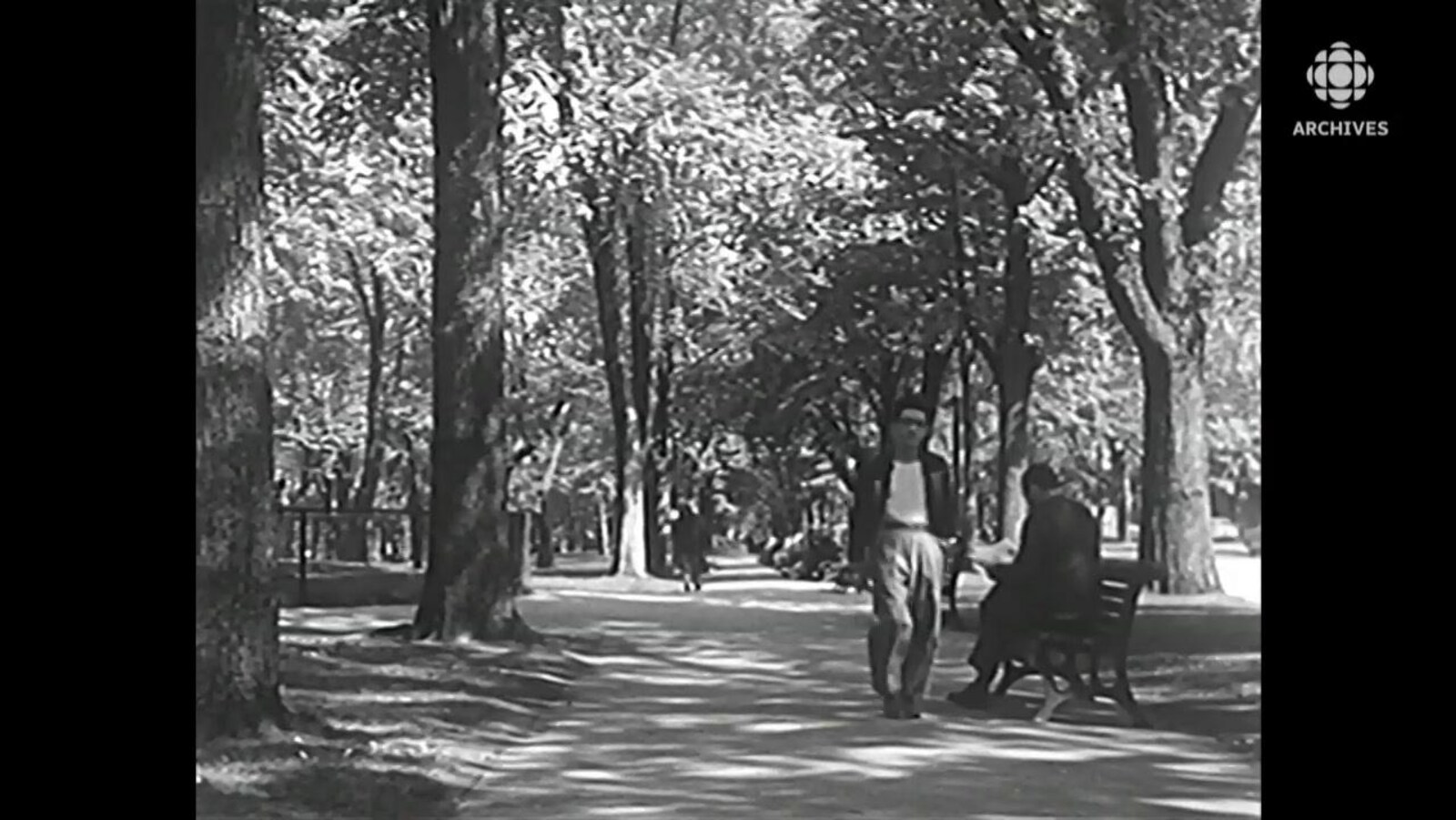 Un homme flâne dans un sentier du parc La Fontaine et circule près d'un autre qui lit son journal sur un banc.