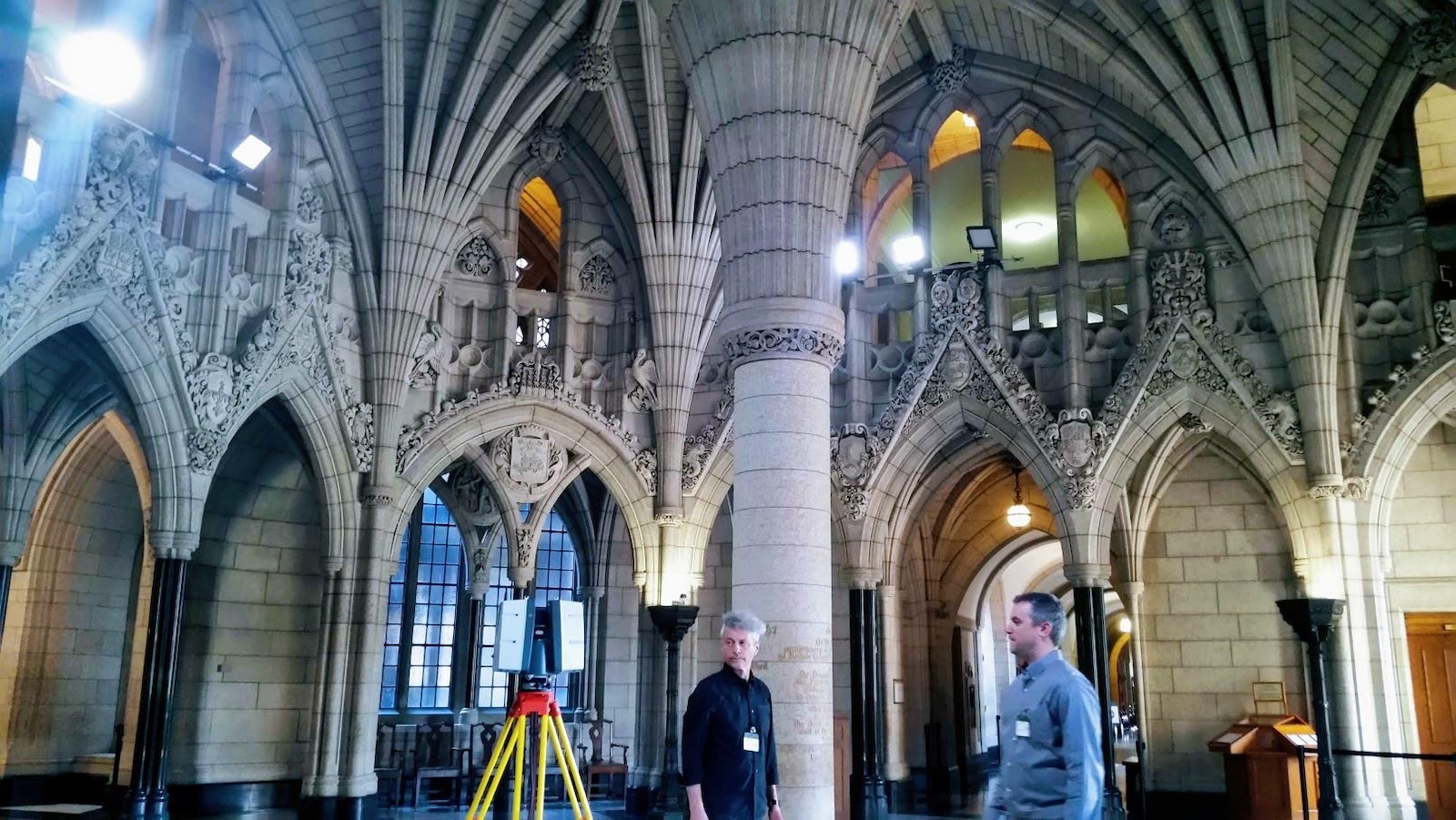 On voit deux hommes dans l'édifice du centre du parlement d'Ottawa, à côté d'eux se trouve un appareil de mesure sur trépied : autour d'eux, on voit les colonnes, les arches, les alcôves.
