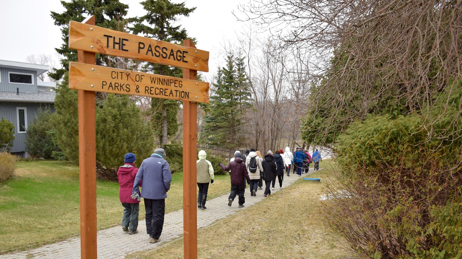 Un groupe de marche suit un trottoir vers la rivière d'où une affiche de la Ville de Winnipeg indique « The Passage ».