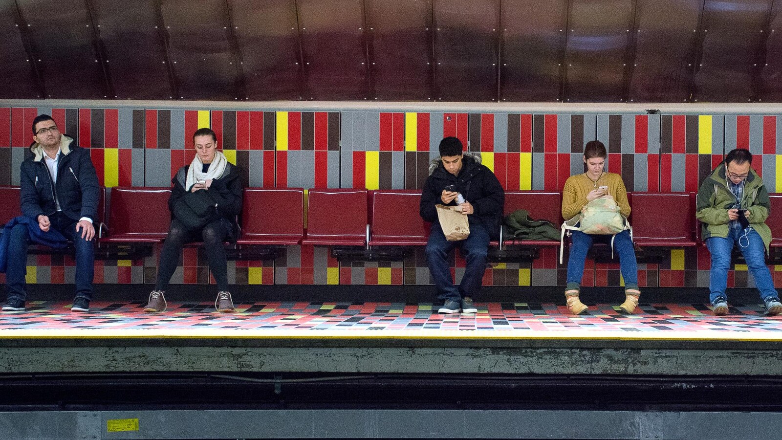 Des gens sont assis sur une banquette, et la plupart d'entre eux regardent leur téléphone.
