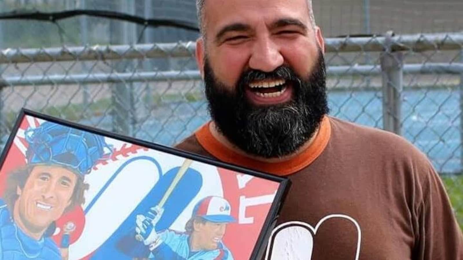 Portrait de l'auteur Patrick Beauséjour souriant devant un terrain de sport et tenant à la main un tableau représentant des joueurs de baseball des Expos.