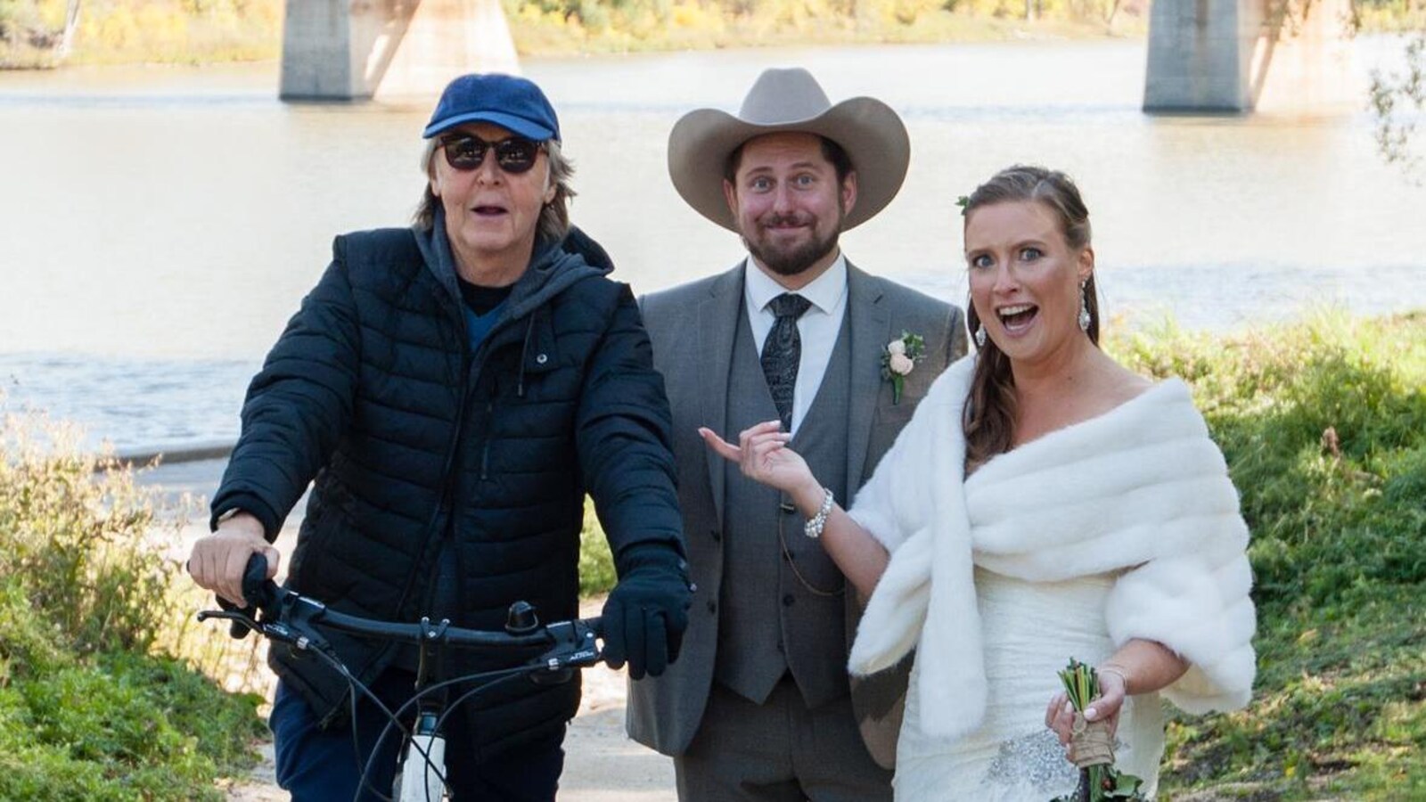 Un homme sur une bicyclette, un homme en habit avec un chapeau de cowboy et une femme en robe de mariée devant une rivière et un pont.