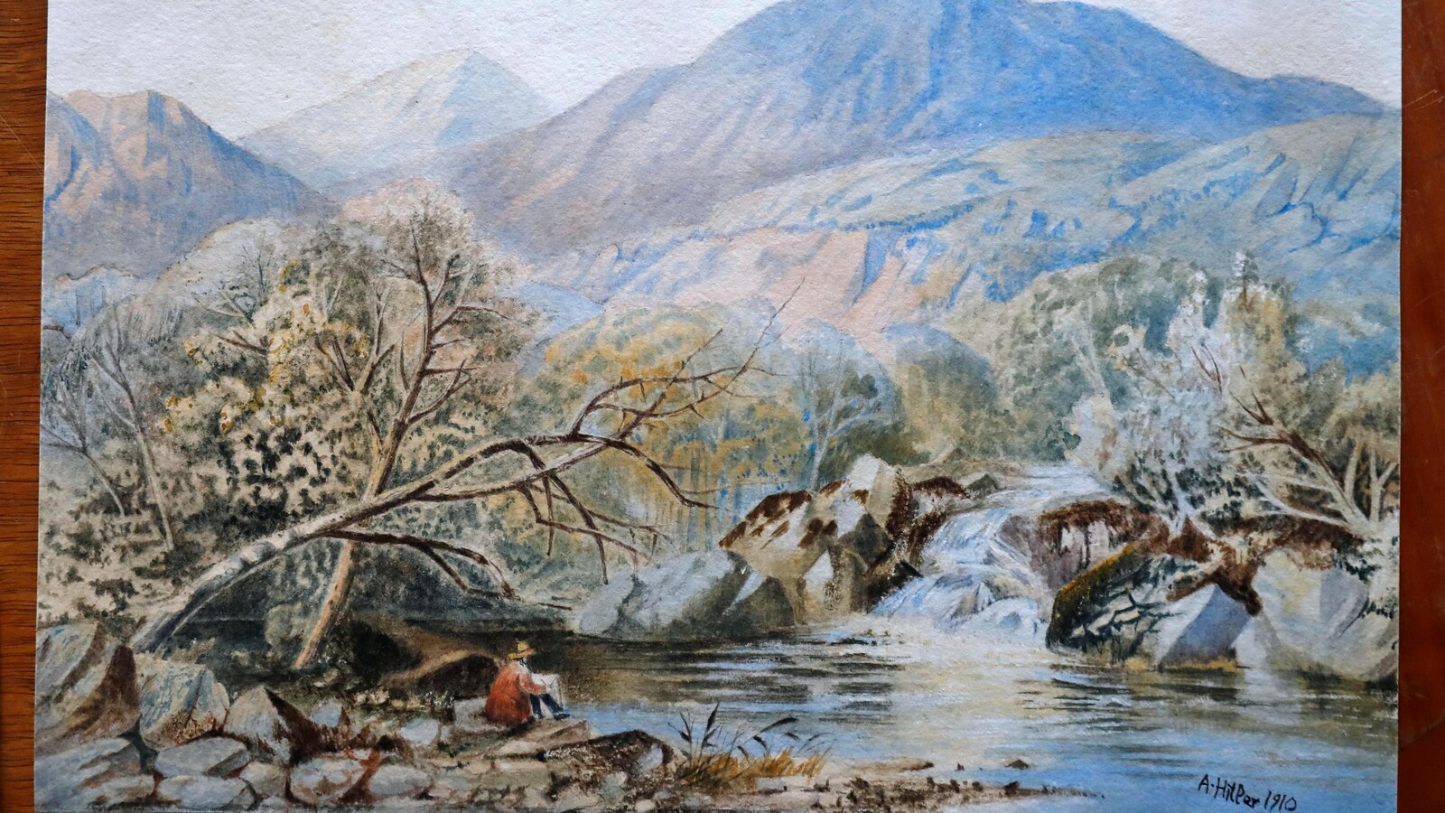 L'oeuvre représente une personne assise au bord d'une rivière, dans un paysage montagneux.