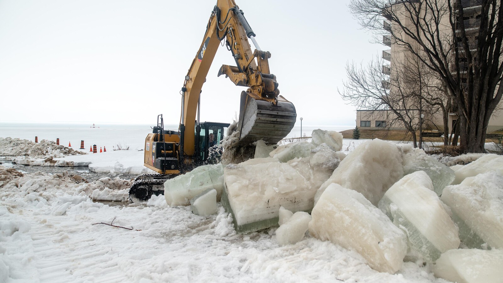 Une pelle mécanique retire d'imposants morceaux de glace d'un ruisseau près d'un immeuble d'habitation.