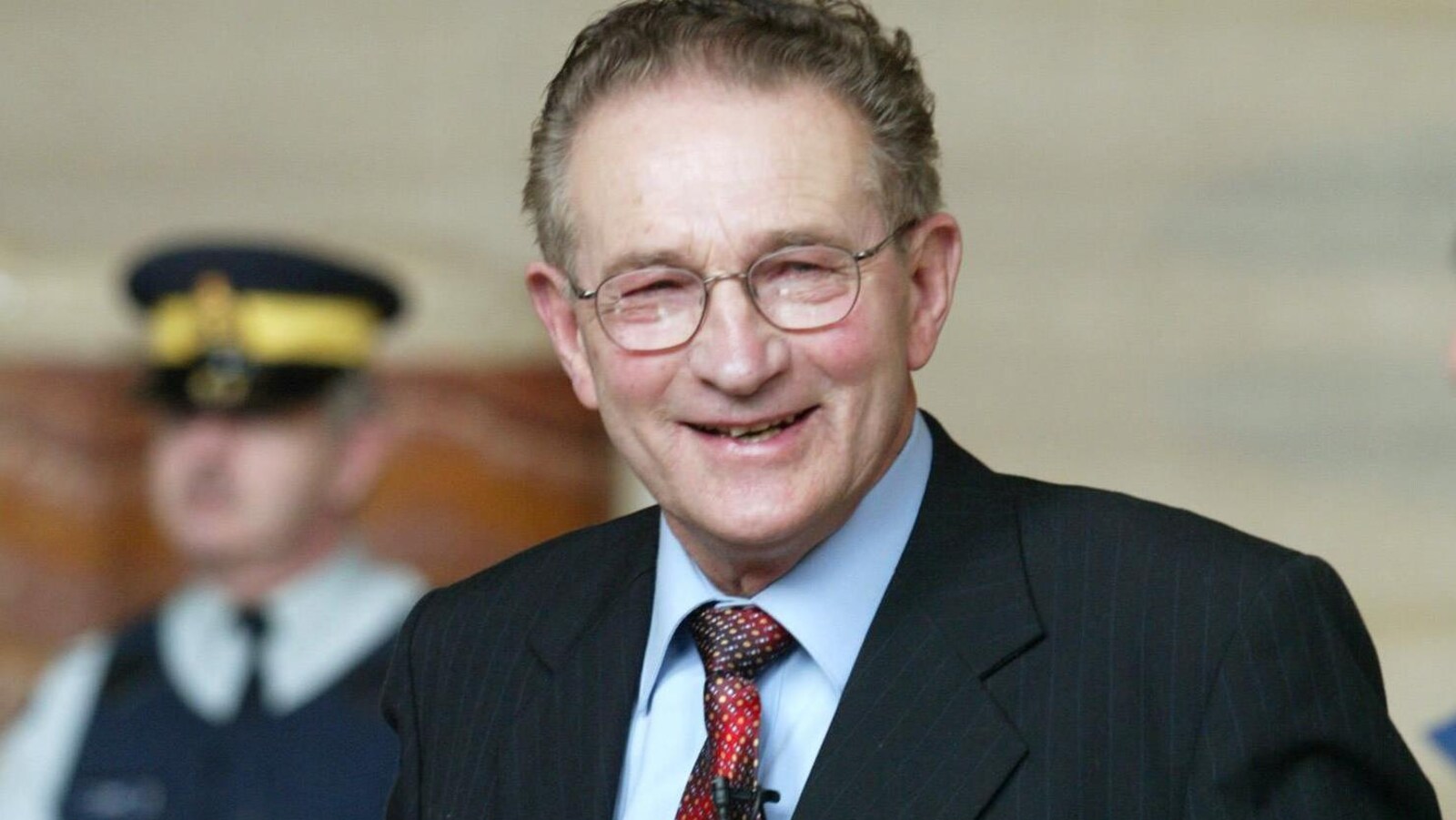Plan serré de Percy Schmeiser en costume cravate qui sourit. En flou en arrière-plan : un agent de la GRC.