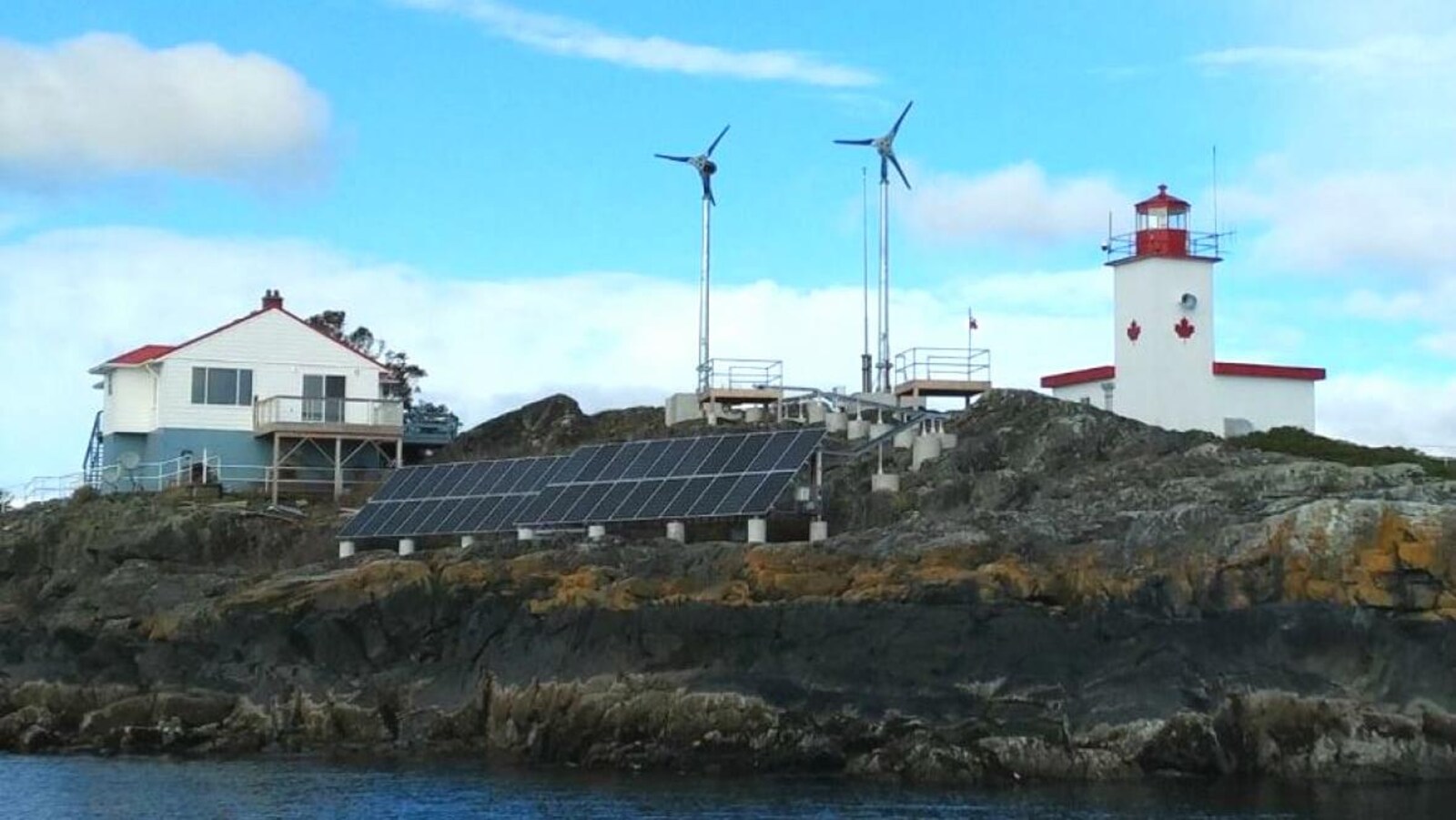 Deux éoliennes surplombent l'île rocailleuse. À leurs pieds se trouvent des panneaux solaires, le phare et la maison du gardien.