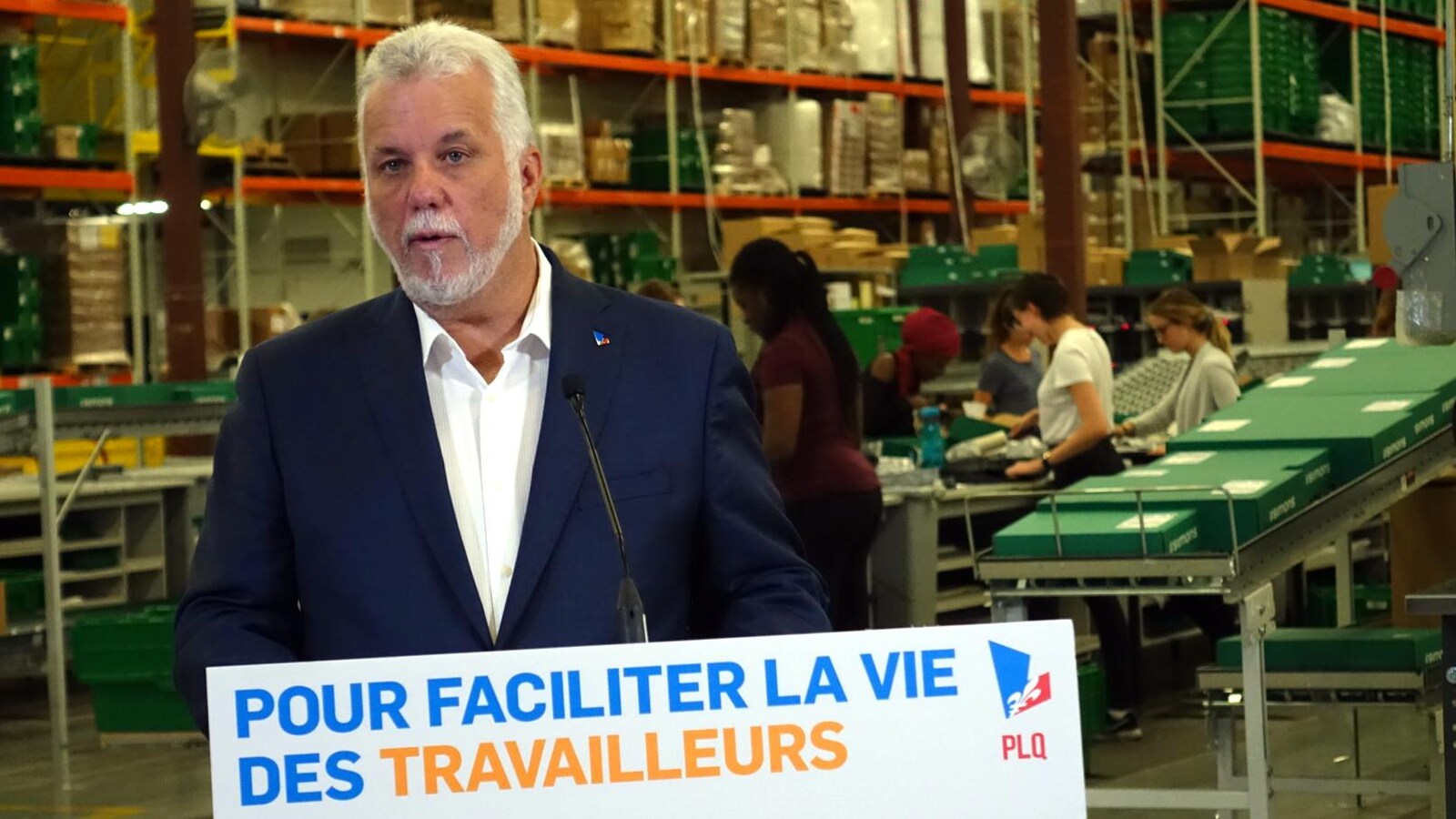 Philippe Couillard fait un point de presse dans le centre de distribution d'un magasin Simons. Des femmes travaillent derrière lui.
