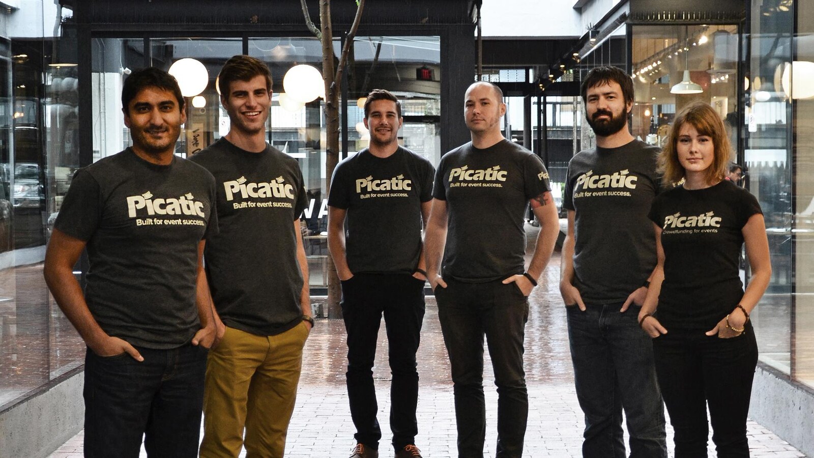 Cinq hommes et une femme disposés en arc de cercle dans le hall d'un bâtiment. Tous portent des T-shirts avec l'inscription «built for event success».