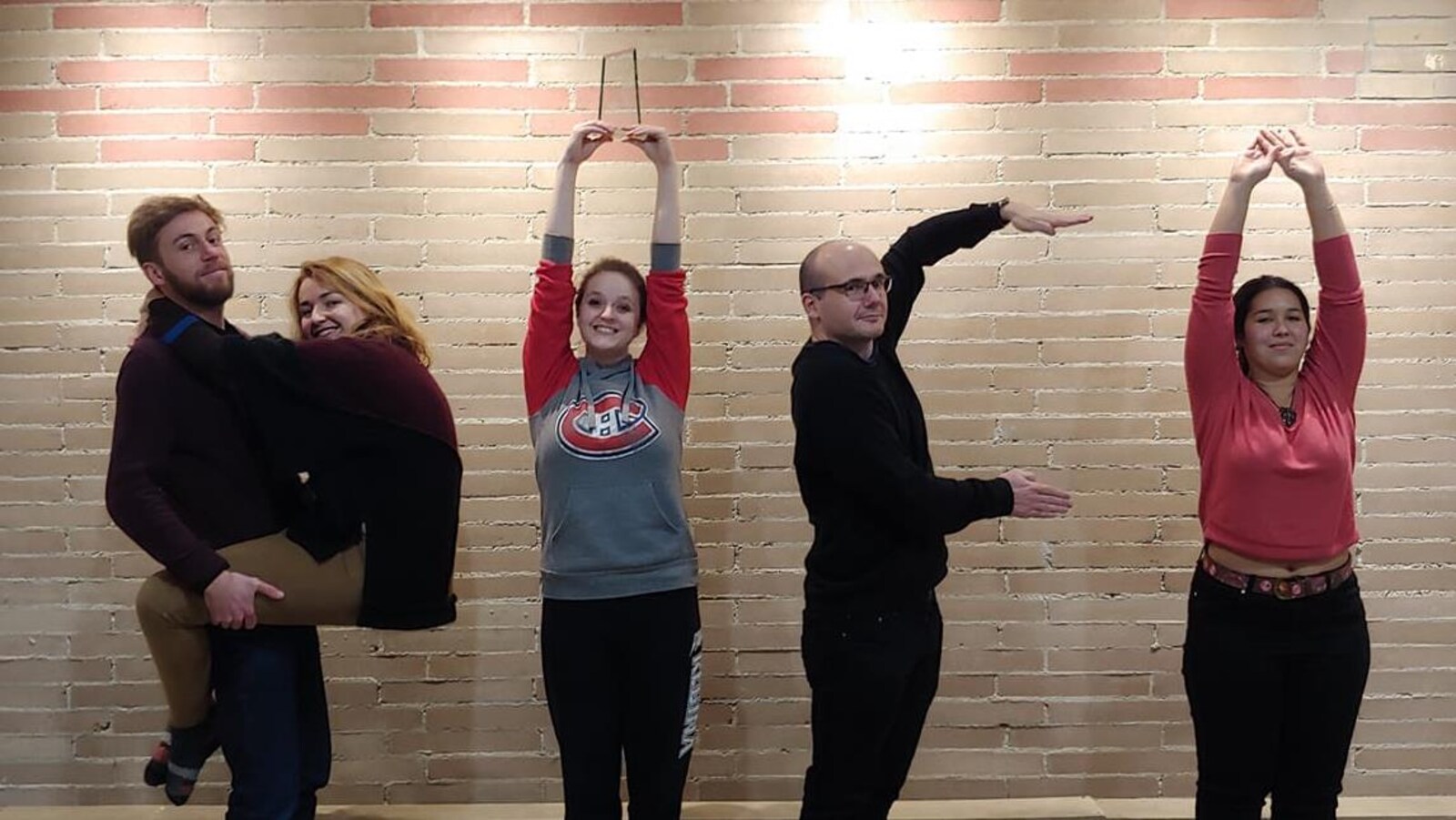 5 personnes qui ont l'air de bien s'amuser prennent la pose pour former les lettres PIF avec un point d'exclamation devant un mur de briques.