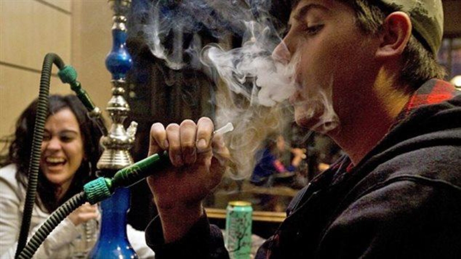 Un jeune homme fume un narguilé dans un bar. Une jeune femme rit en arrière plan.