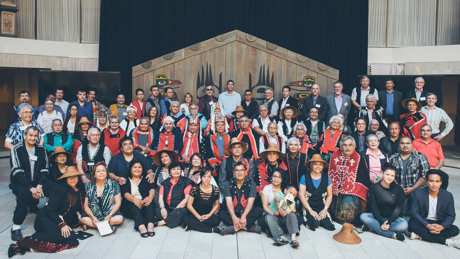 Environ 80 membres de Premières Nations, certains avec des habits ou chapeaux traditionnels, dont un enfant et plusieurs personnes âgées posent pour une photographie de groupe.