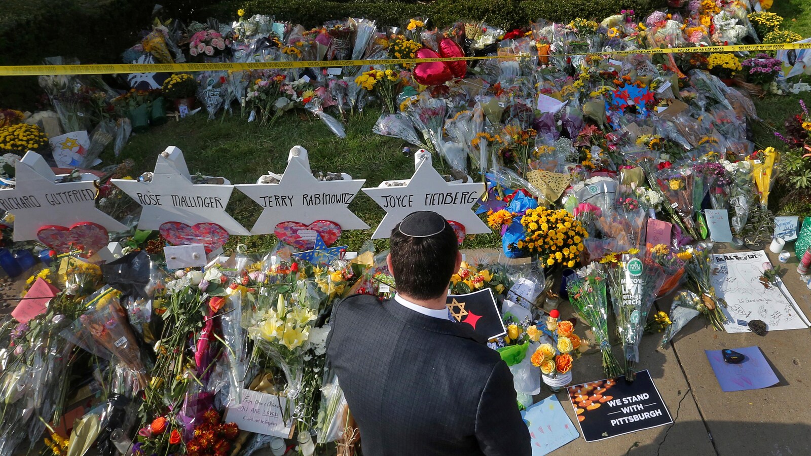 Un homme portant une kippa est vu de dos. Il est debout devant des dizaines de bouquets de fleurs et des étoiles en carton au nom des victimes.