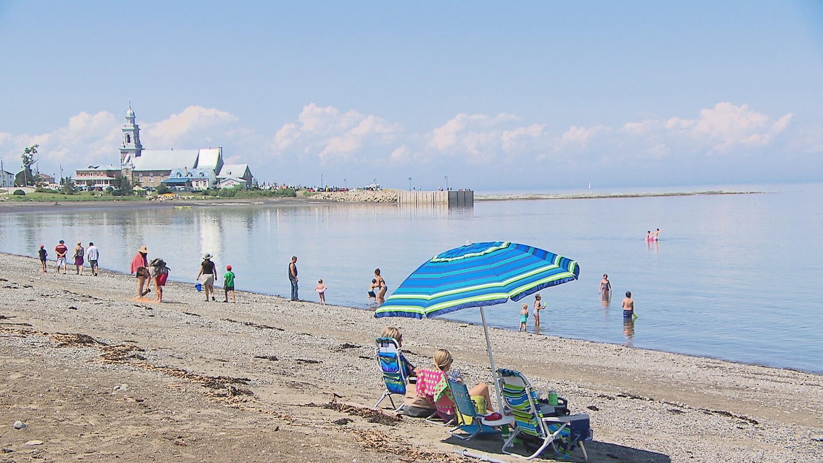 Une dizaine de personnes profitent de la plage, des enfants se baignent tandis que deux femmes se reposent sous un parasol bleu.