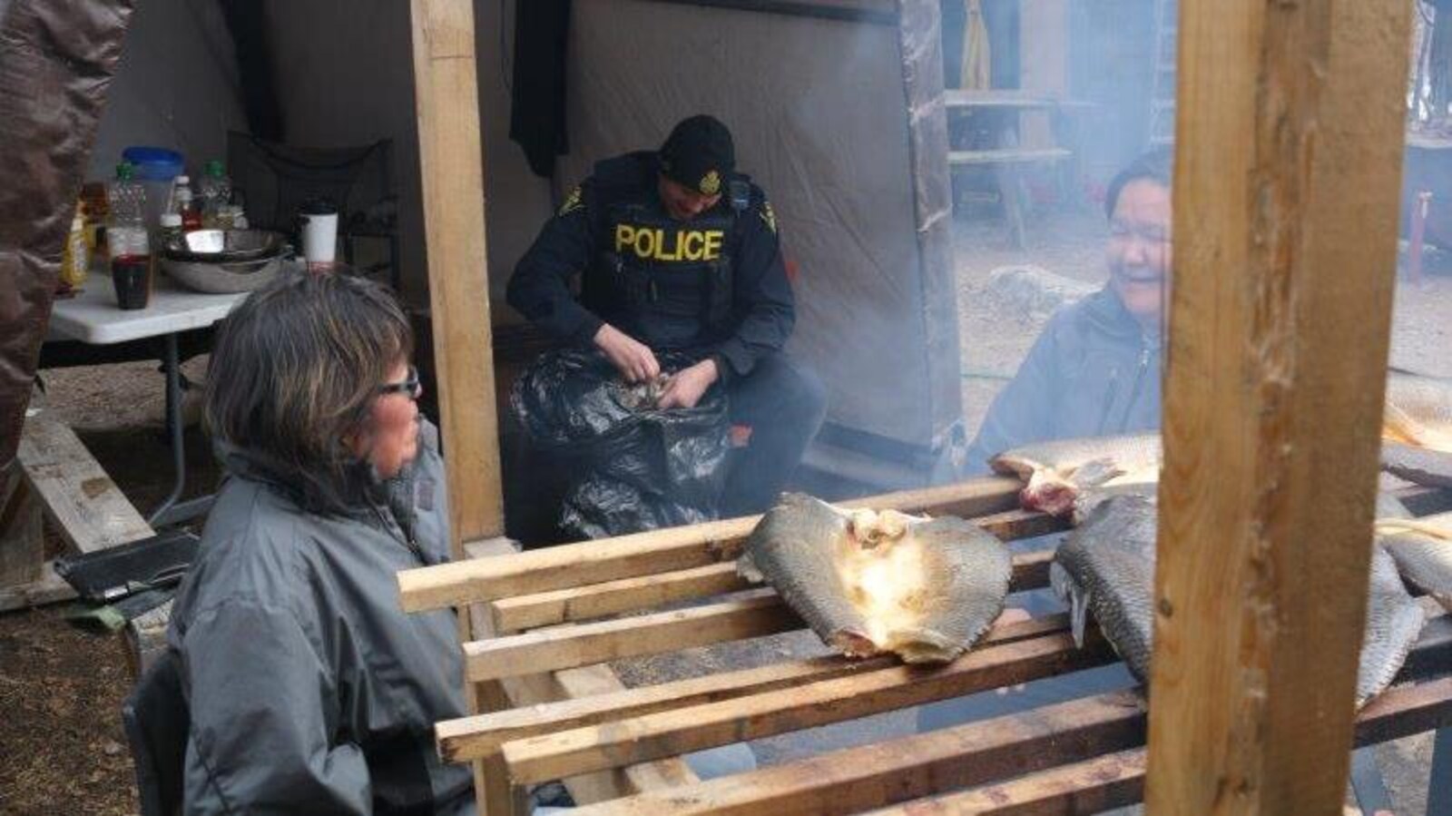 Des femmes discutent devant du poisson en train de fumer avec un policier derrière.