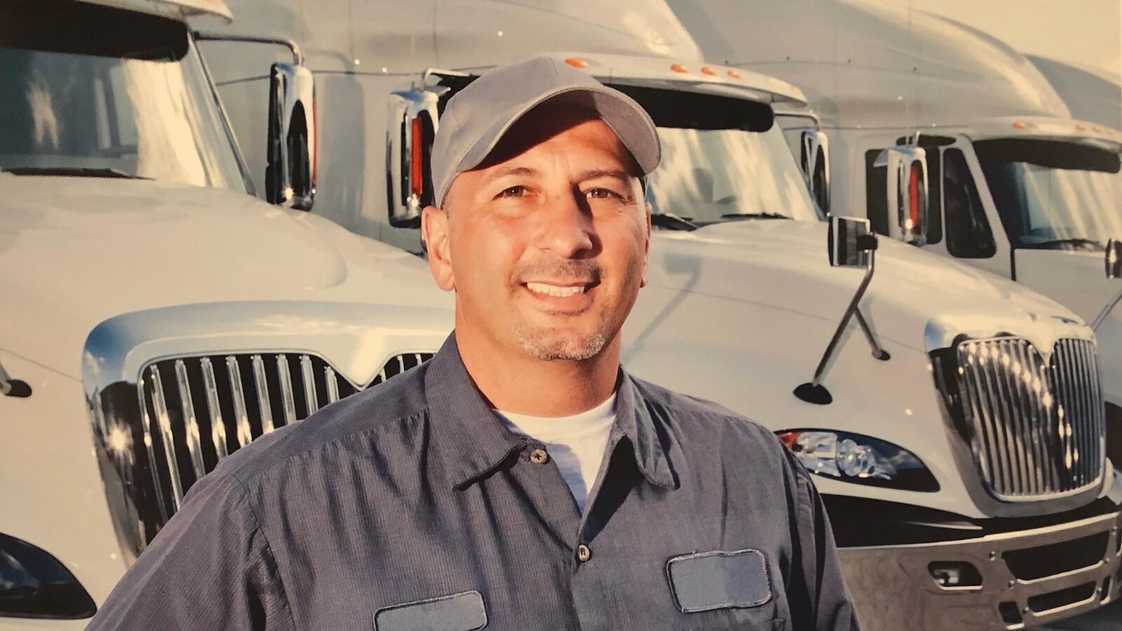Photo d'un camionneur portant une chemise et une casquette bleues devant des poids lourds stationnés.