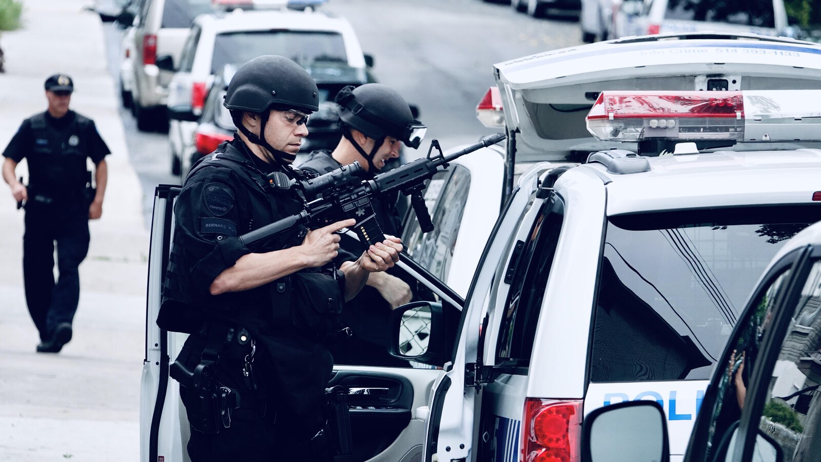 Deux policiers, casqués, dont l'un au premier plan est armée d'une arme de type mitraillette. Ils sont tout près d'un véhicule de police, à la droite de l'image.